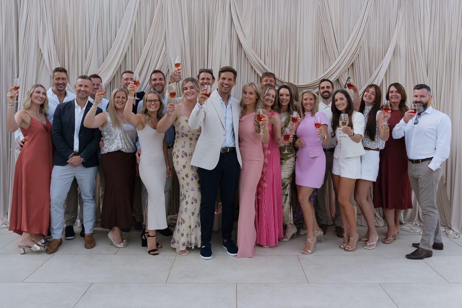 Large group of elegantly dressed guests celebrating at a wedding reception, raising glasses for a toast outdoors.