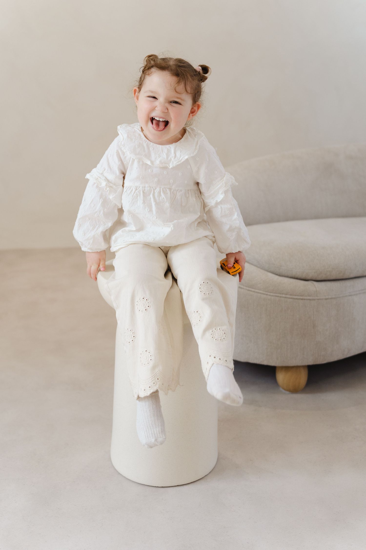 Baby in white christening gown sitting on white pedestal against neutral background.