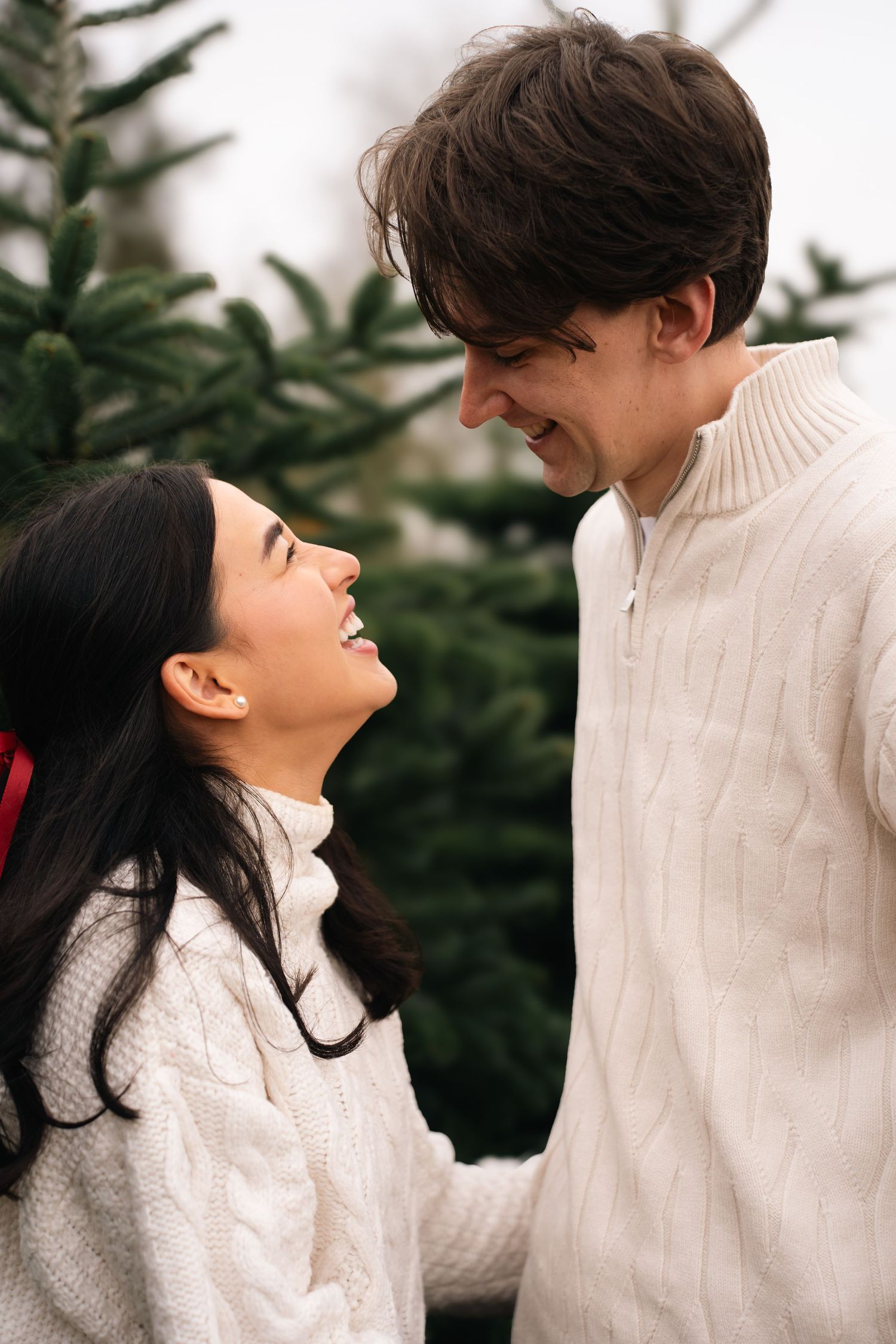 A couple in cozy white sweaters share tender moments against a backdrop of evergreen trees in winter.