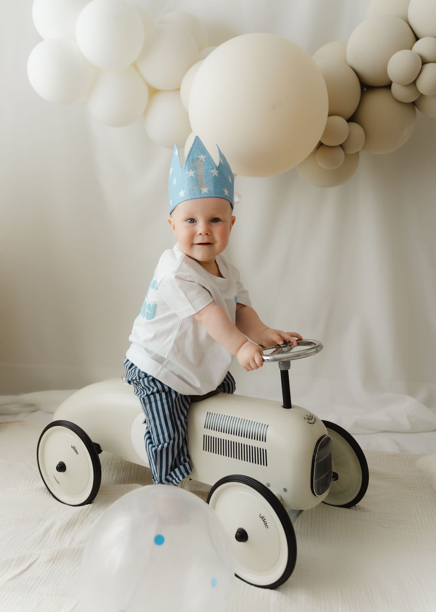 A vintage-style white toy car with balloon decorations in the background makes for a charming first birthday photo setting.