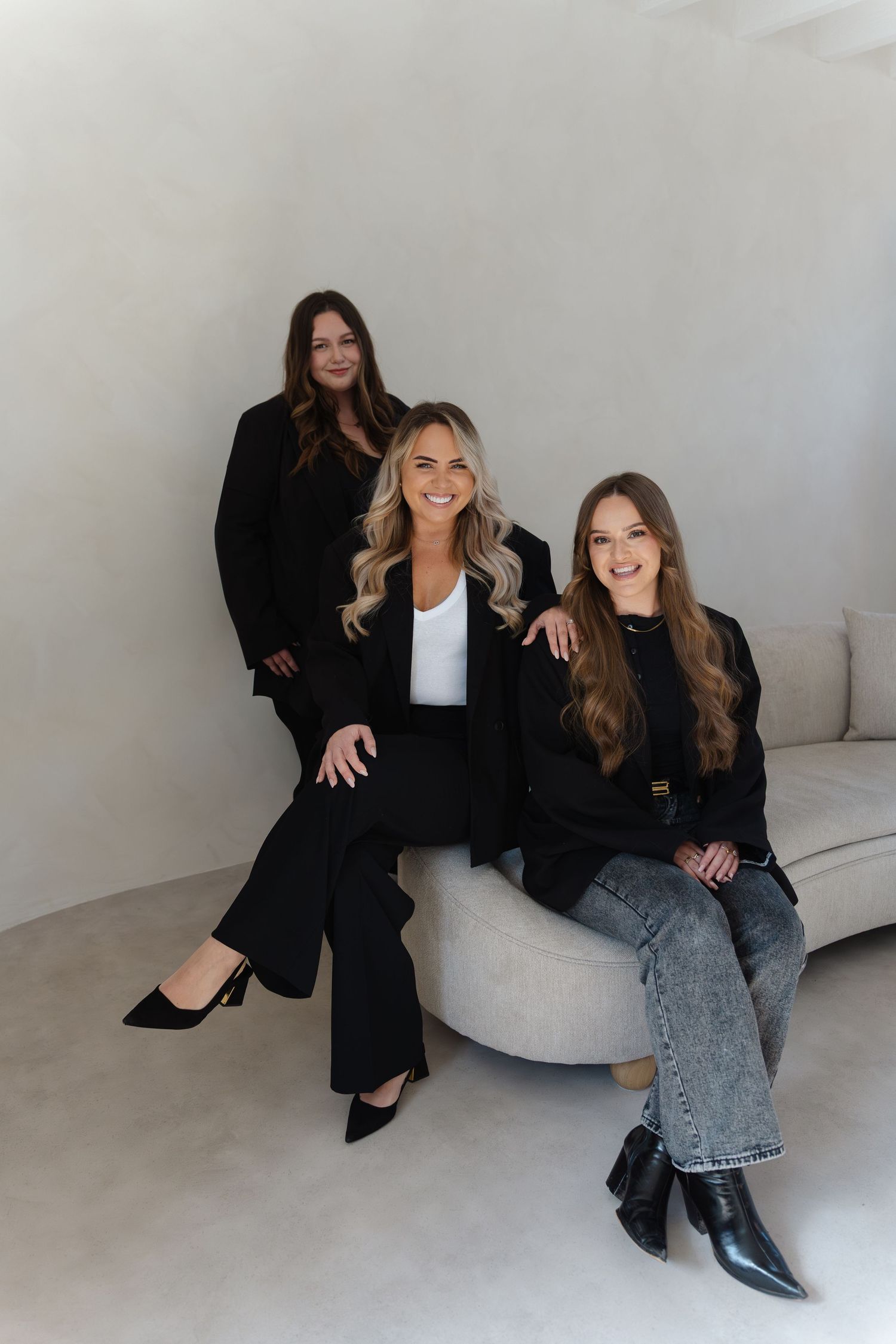 Two people sitting on a white couch in business attire pose for professional photos in a minimalist studio setting.