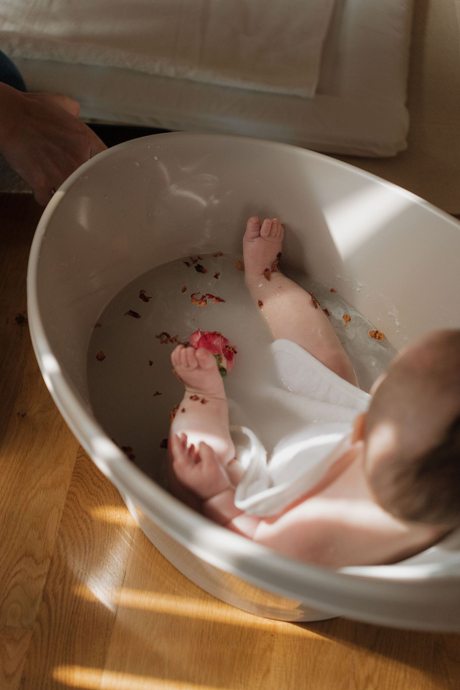 Baby enjoying a bath in a white oval tub on a wooden floor, with colorful marks on skin in natural light.