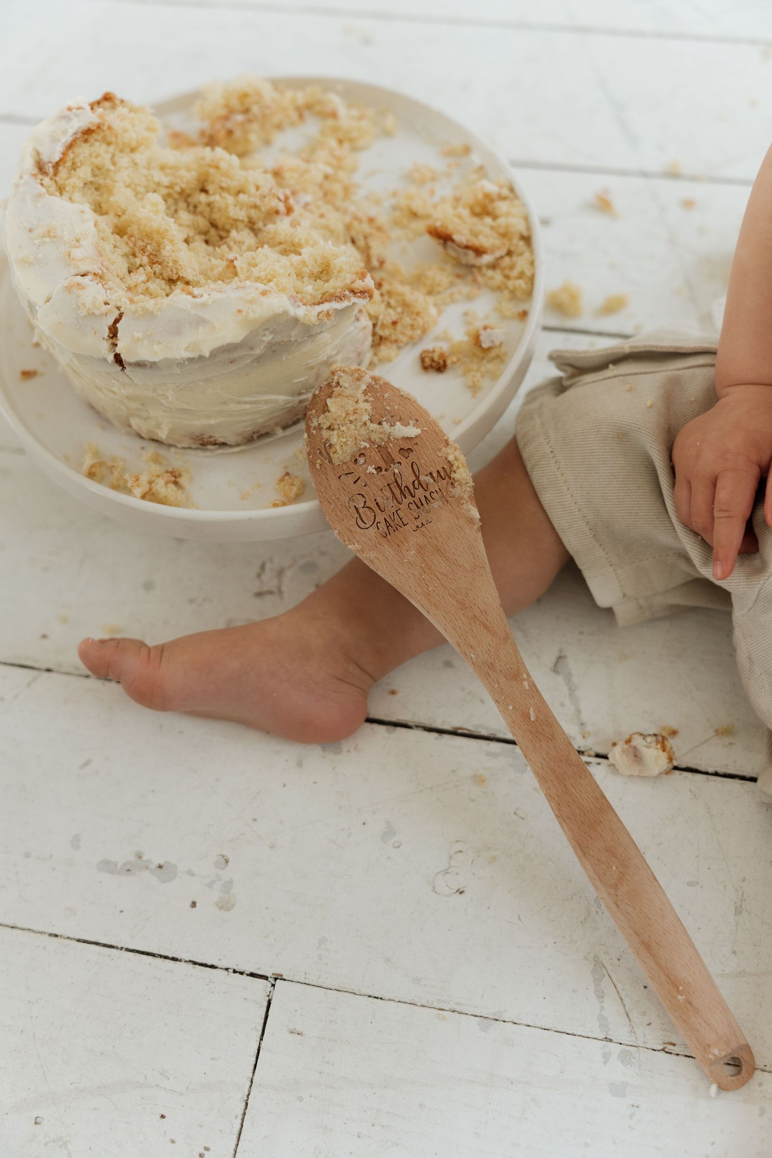 Child sitting on white floor beside a white frosted cake with crumble topping and an engraved wooden spoon.