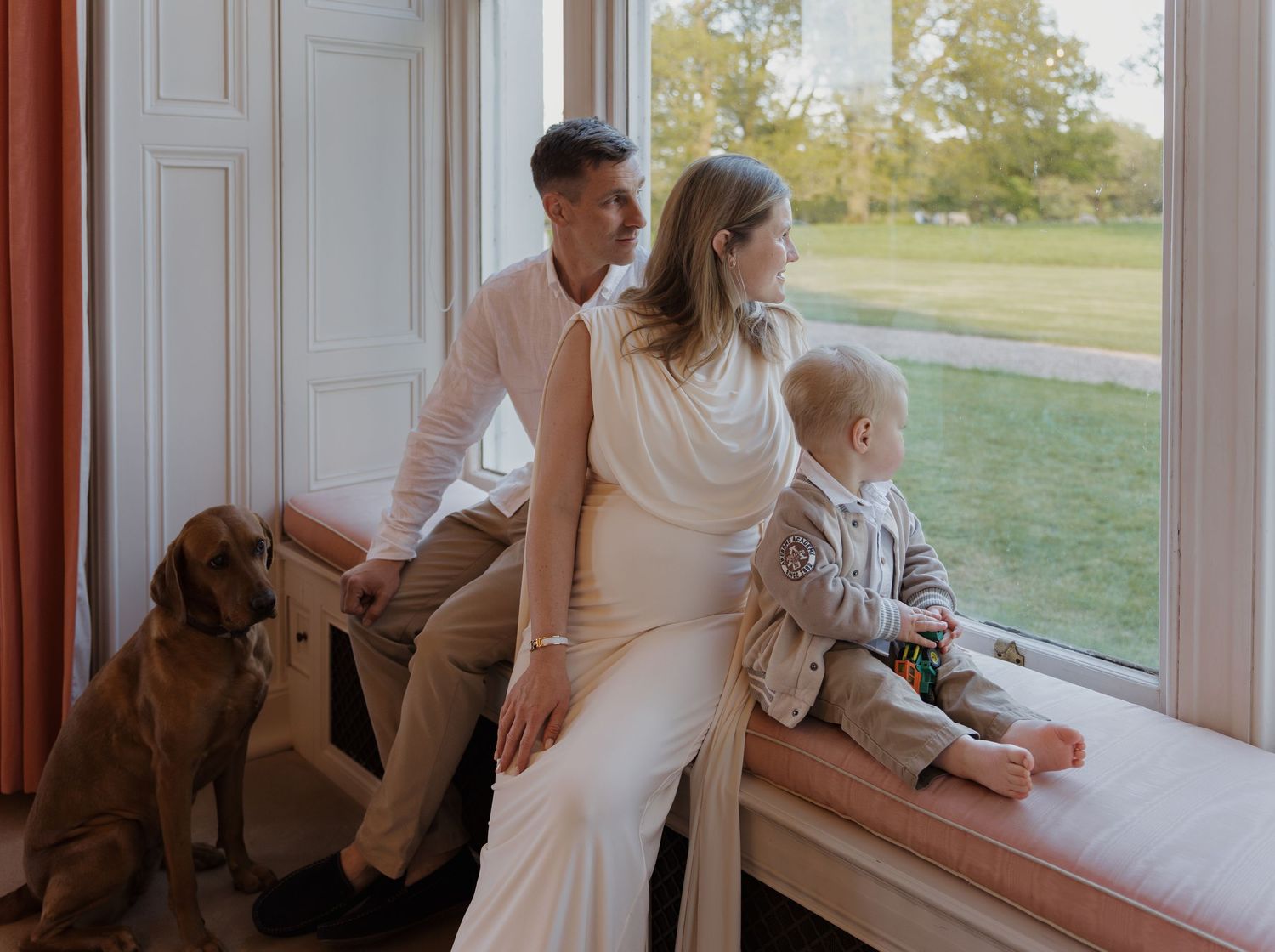 Family posing by large window with dog, woman in white dress, man in white shirt, toddler on windowsill, green garden view.