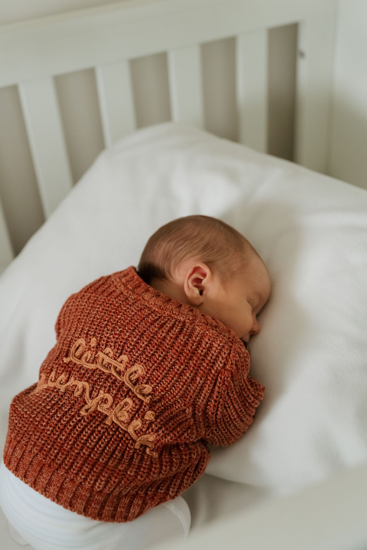 Baby sleeping peacefully in white crib wearing cozy rust-colored knit sweater. Documentary newborn photography in Derbyshire.
