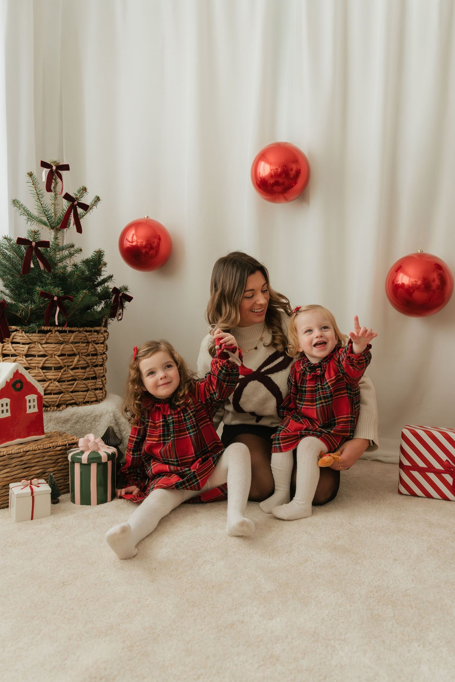 A mother and two young daughters in matching red plaid dresses pose by a Christmas tree with red balloon decorations.