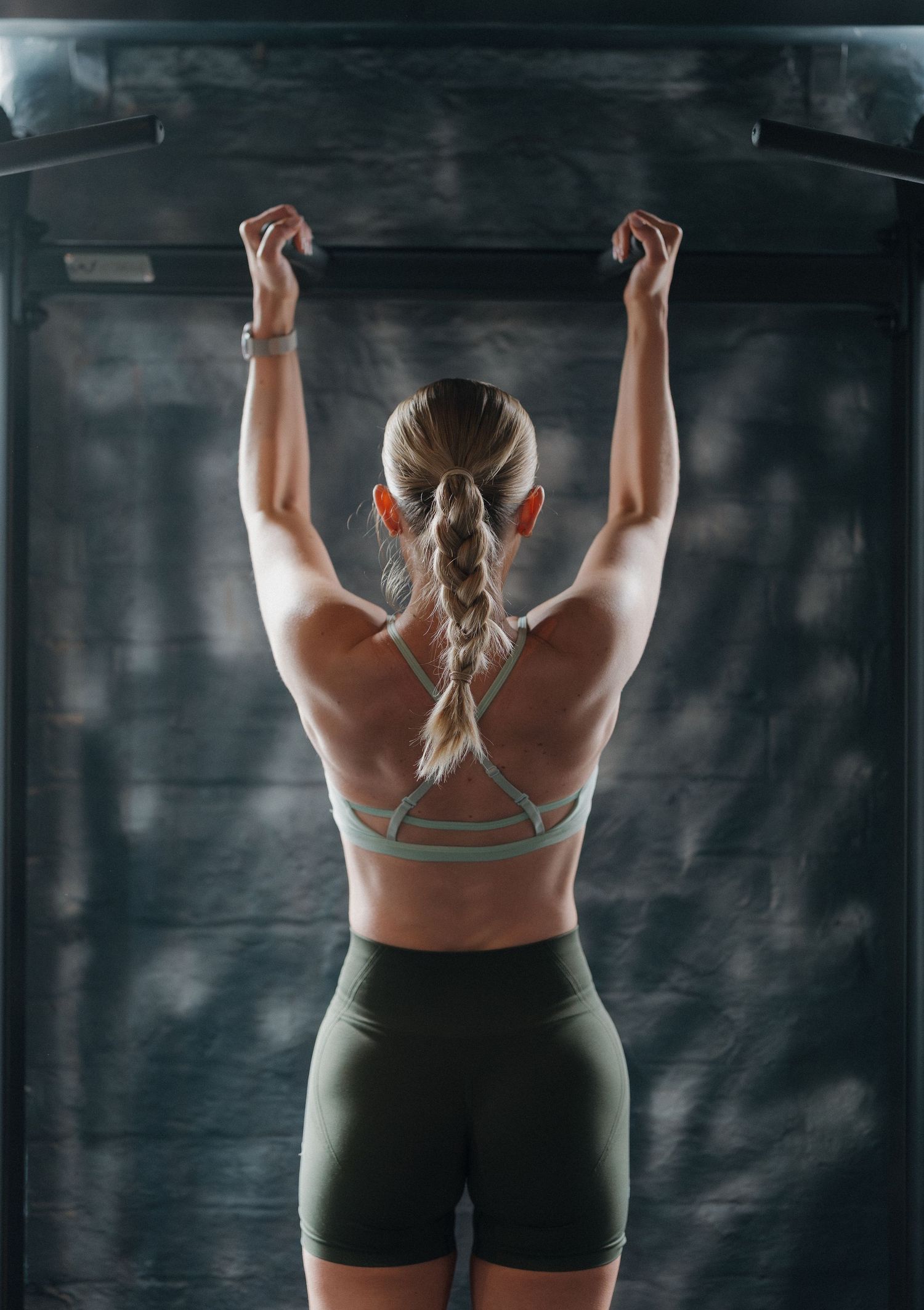 A fit woman in athletic wear hangs from a pull-up bar, showcasing her toned back muscles in a gym setting.