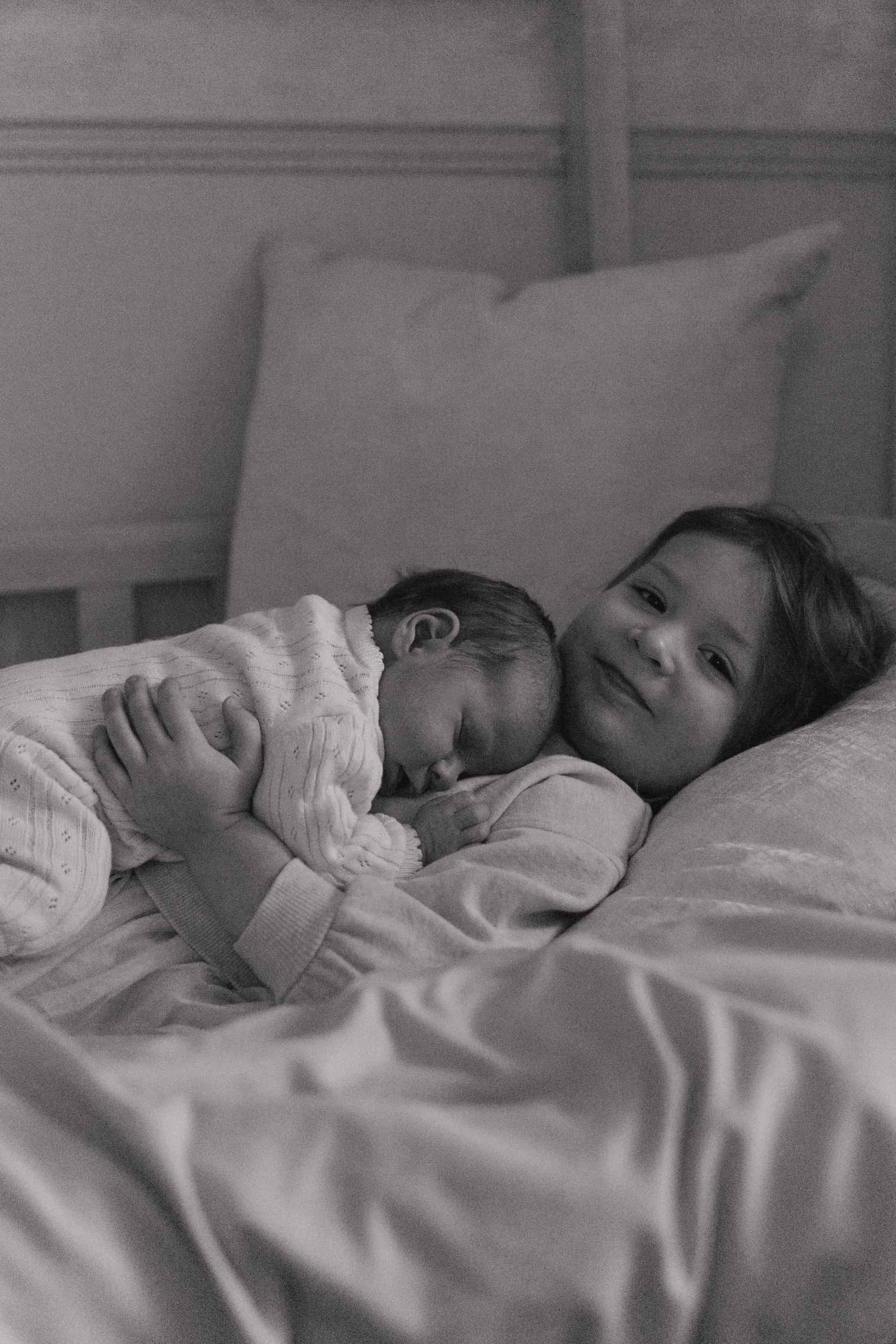 Black and white photo of a young child tenderly holding a sleeping newborn baby on a white bed.