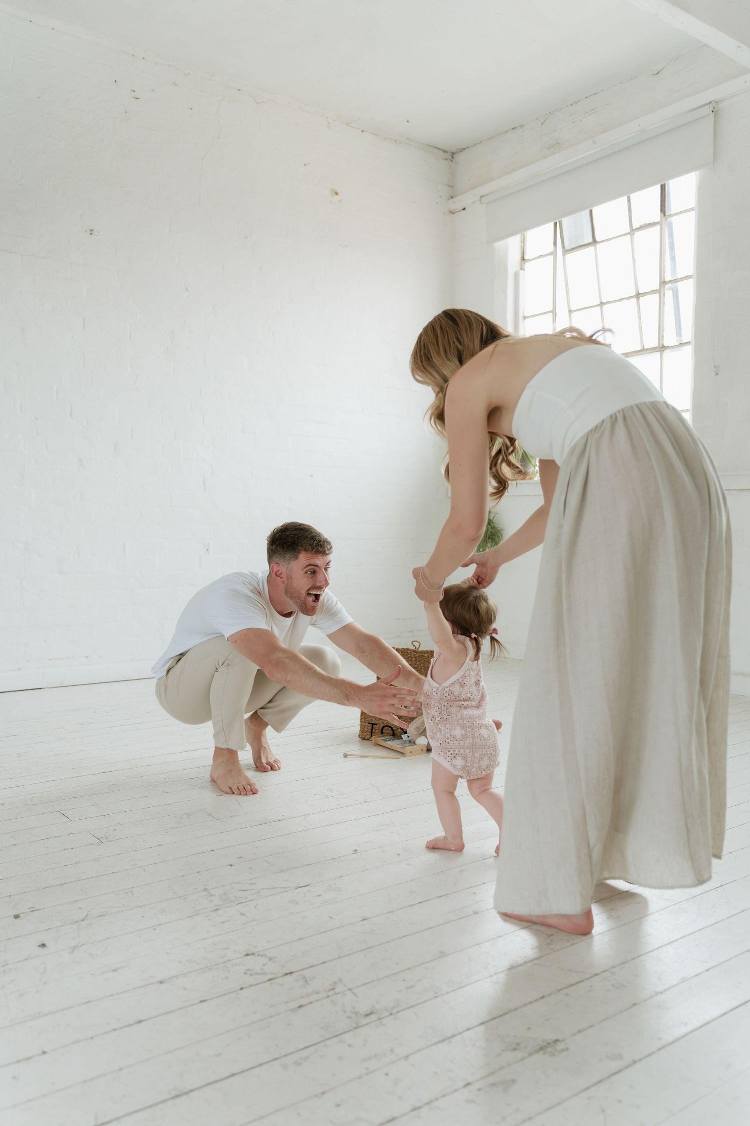Young family with parents encouraging their toddler's first steps in a bright white room.