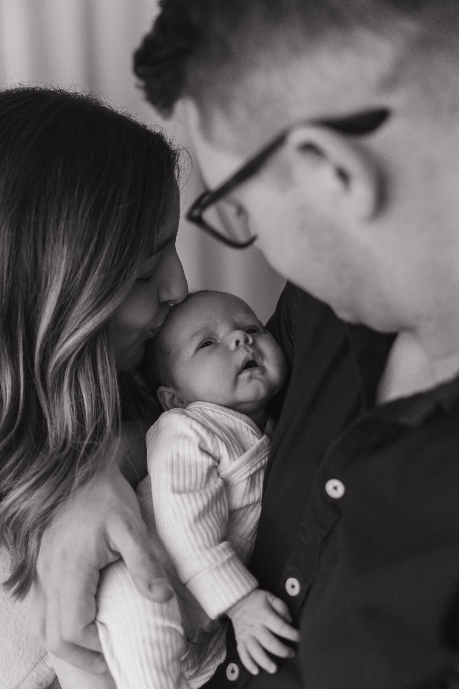Black and white intimate portrait of parents with their sleeping newborn baby in a ribbed white onesie. Documentary newborn photography.
