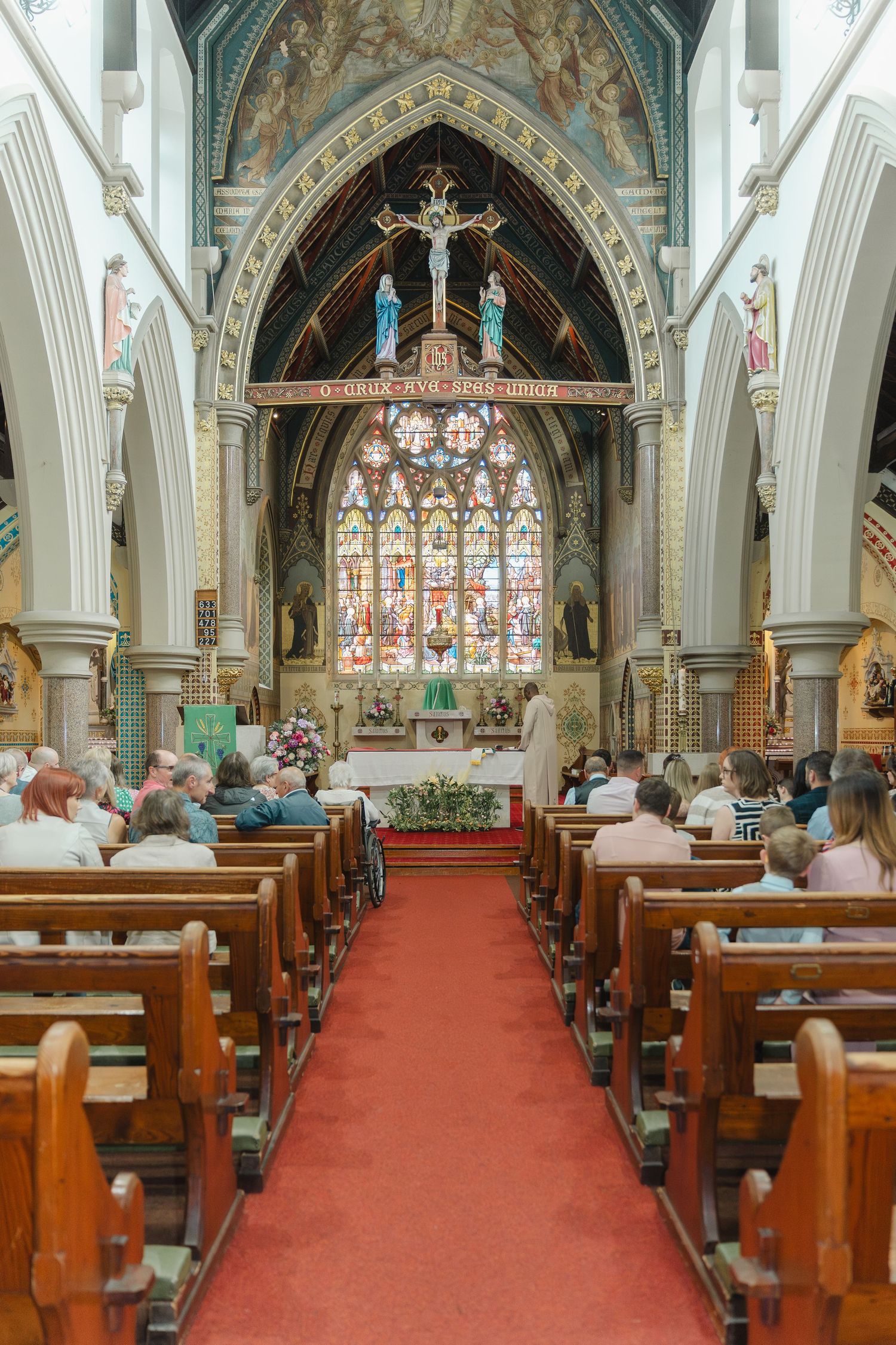 Interior panorama of a Gothic church with red carpet aisle, wooden pews and ornate stained glass windows at the altar.