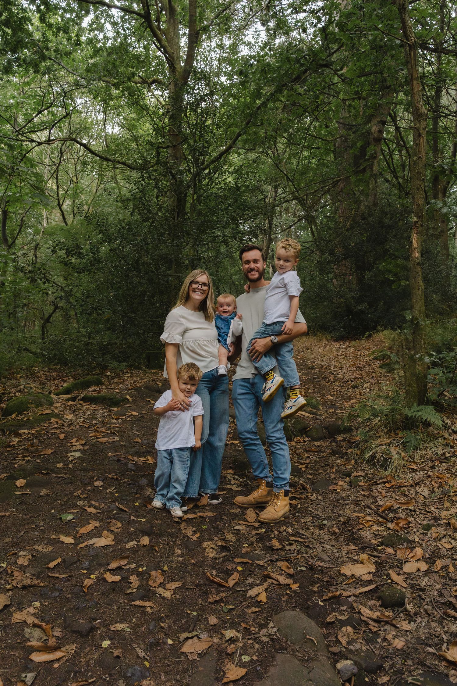 Family wearing coordinated denim and grey outfits poses together on a wooded trail covered in fallen leaves.