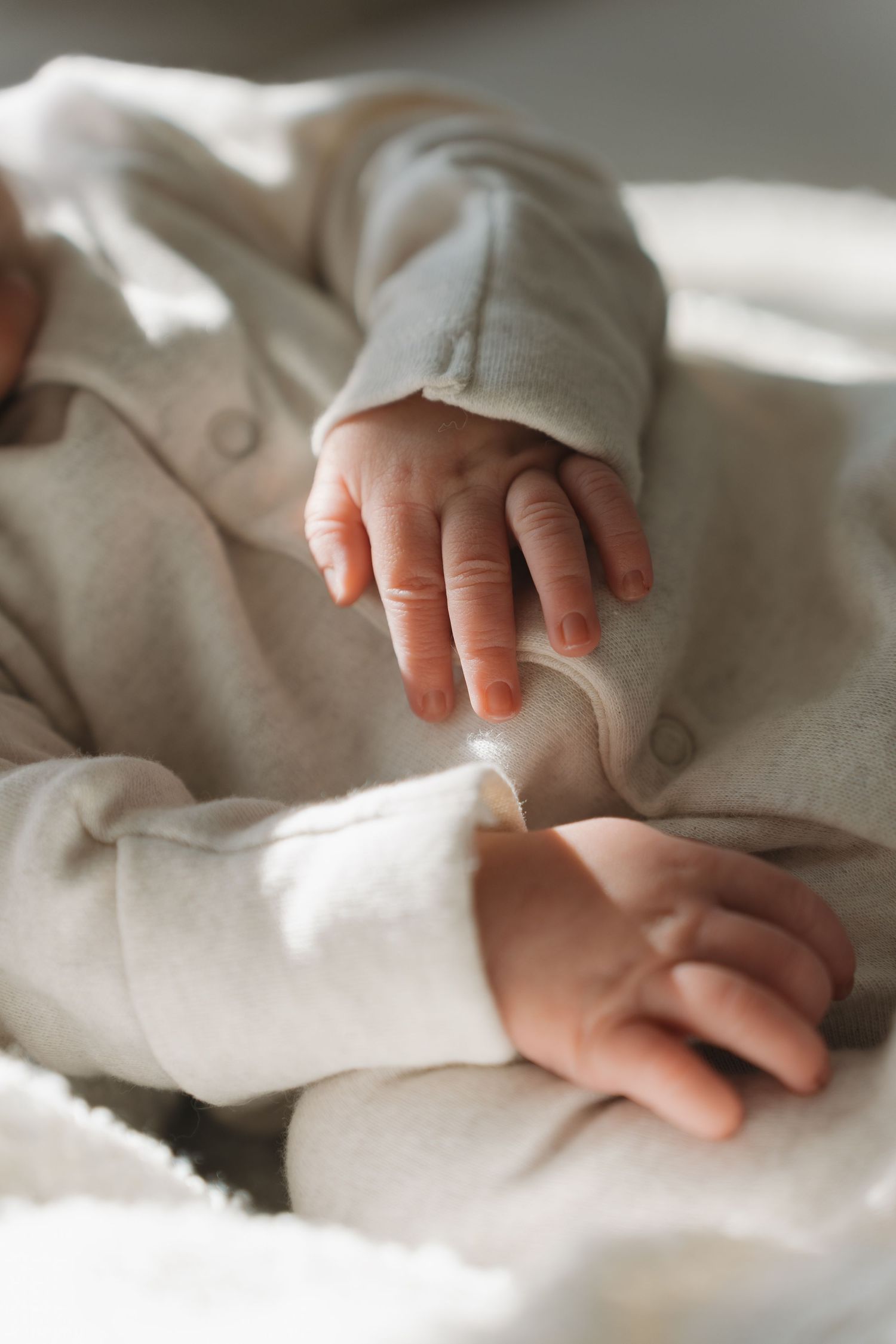 Close-up of a newborn baby's tiny hands resting on a soft white blanket, wearing a cozy light gray onesie. Home Newborn Photography Derby.