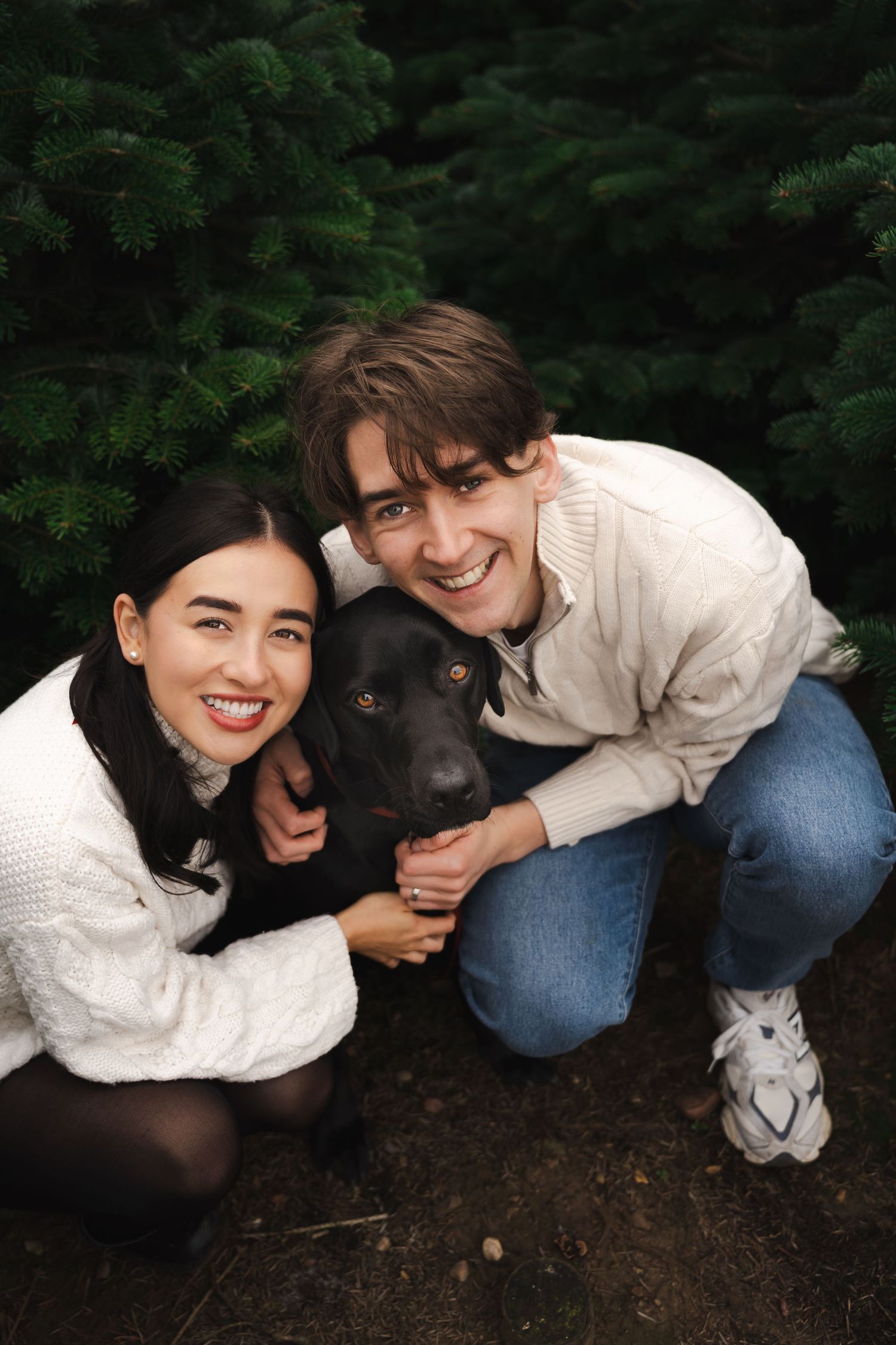 A smiling couple in white sweaters poses outdoors with their black Labrador dog in front of lush green pine trees.