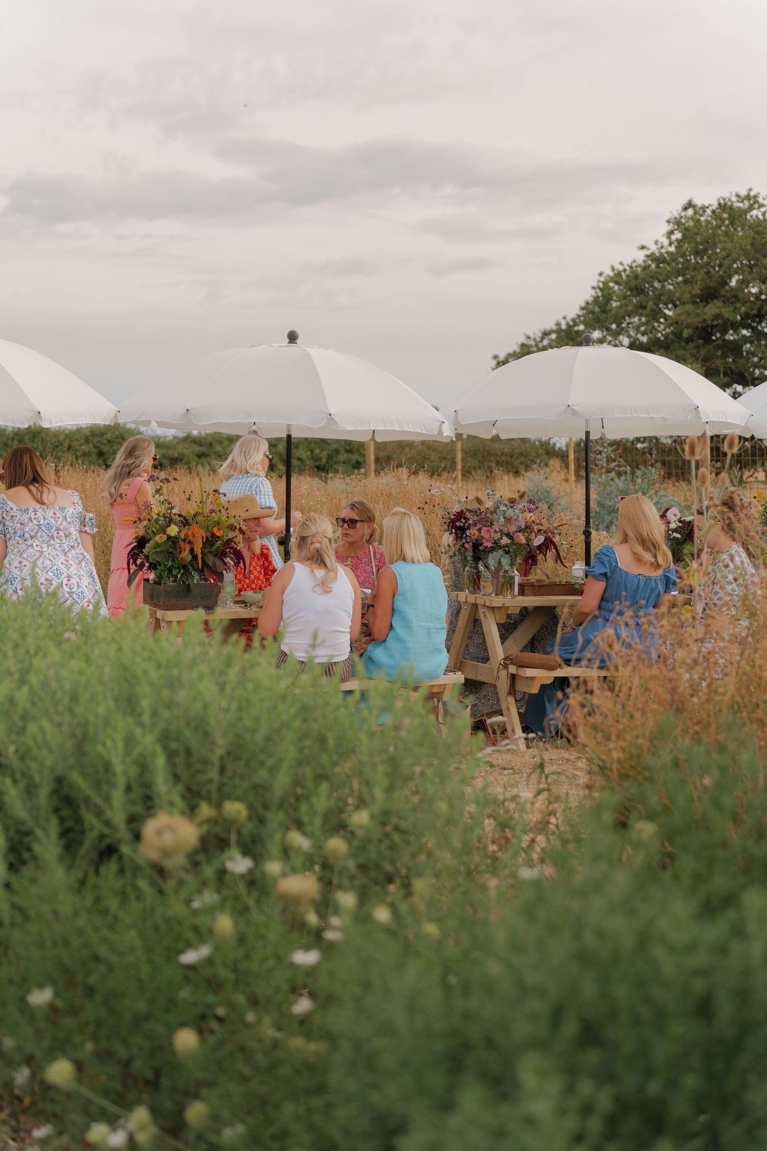 Women gather outdoors at rustic wooden tables with white umbrellas and floral arrangements in a scenic wildflower field.
