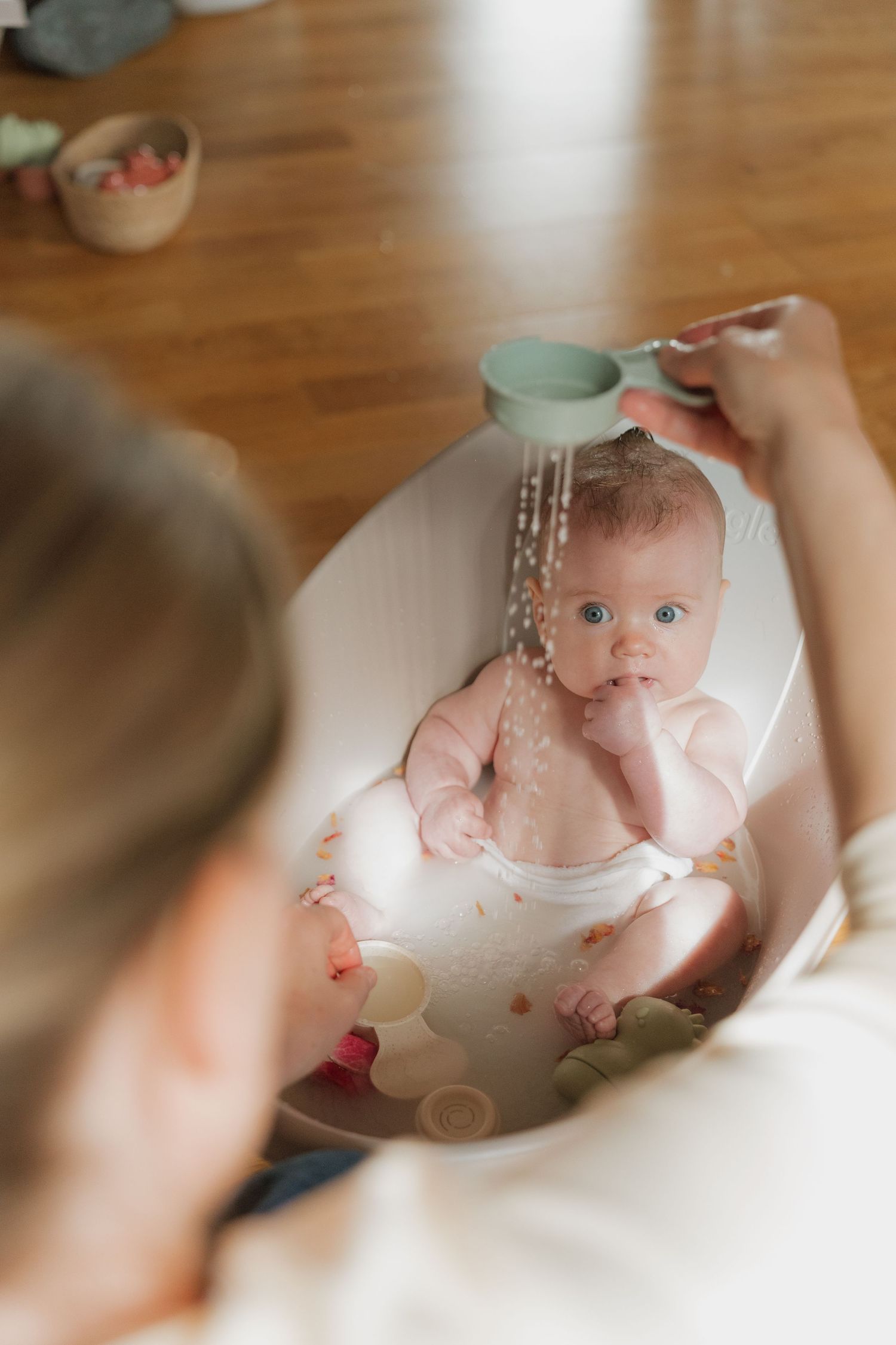 A parent gently rinses a wide-eyed baby during bath time using a small cup in a white infant tub.
