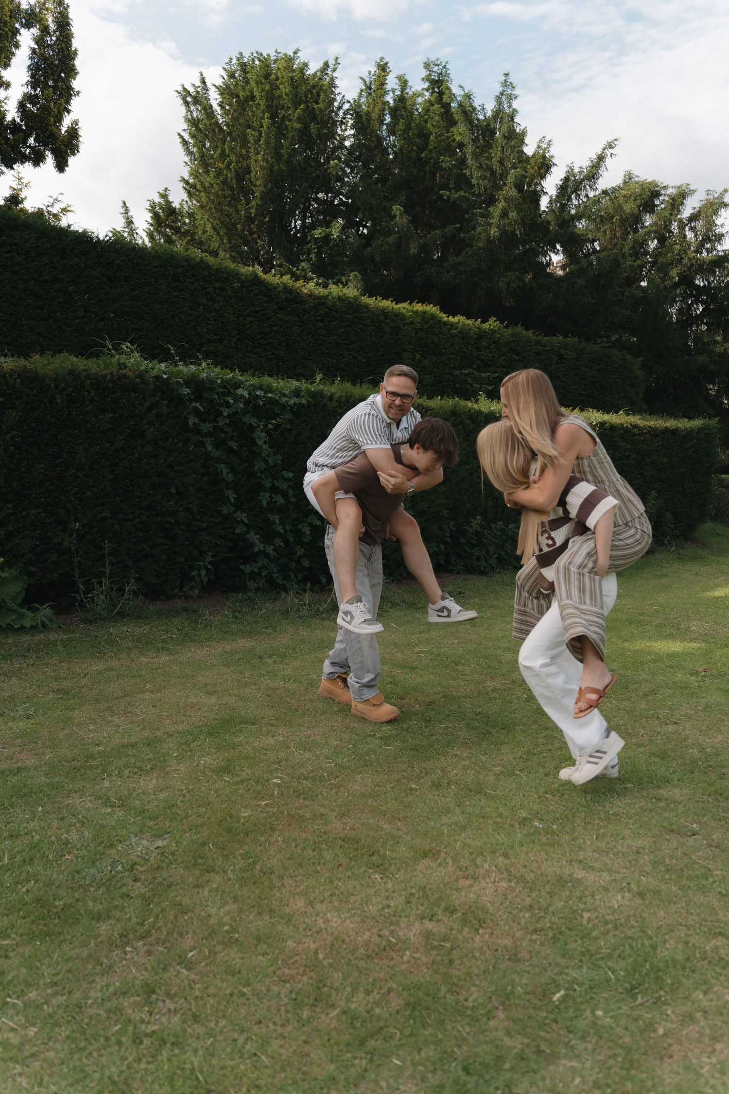Two couples playing piggyback rides on a green lawn with tall hedges and trees in the background.