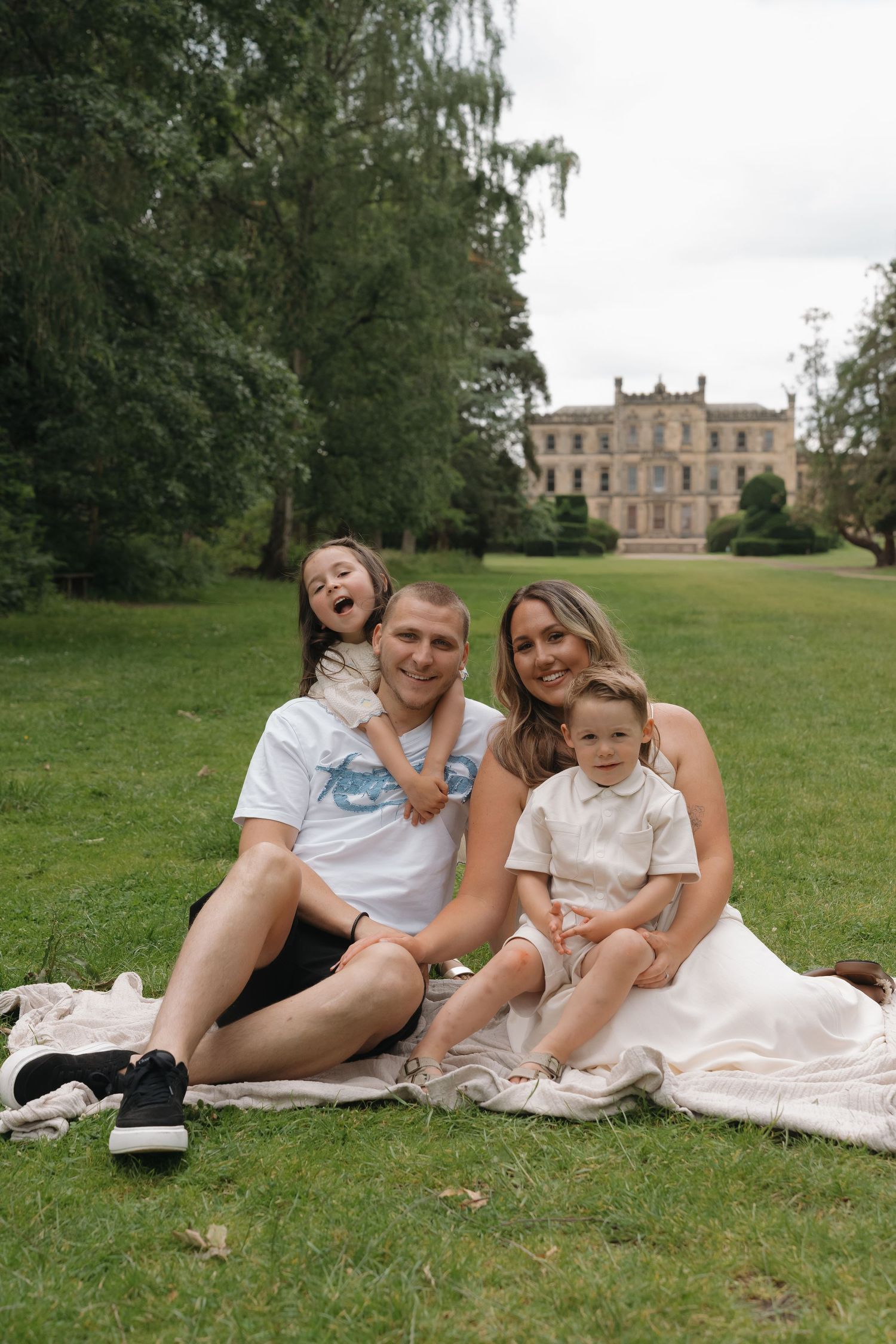 Happy family of four sitting on a blanket in a lush green park with a historic mansion in the background.