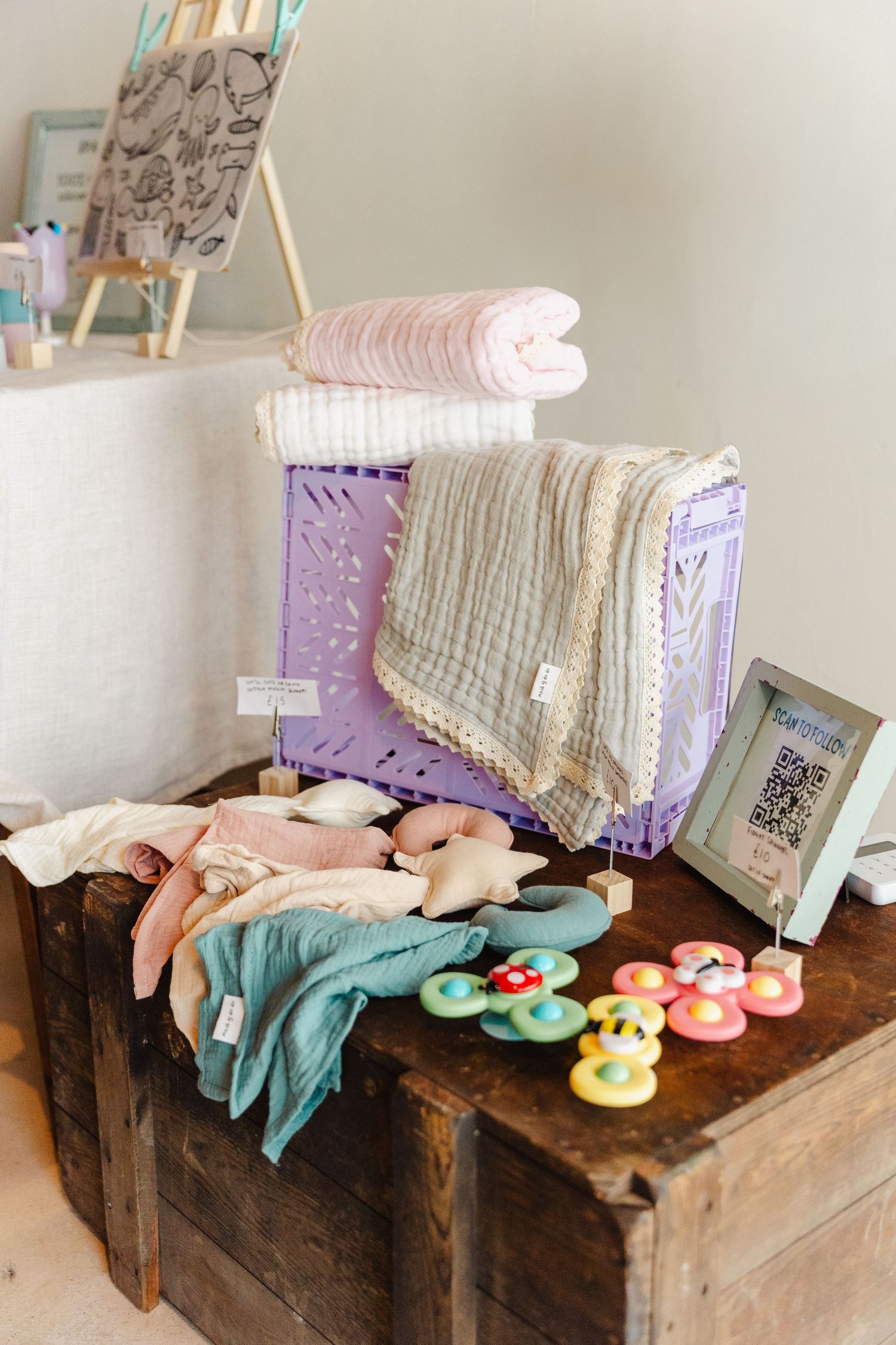 Rustic wooden bench display with folded blankets, plush toys, and baby accessories in soft pastel colors.
