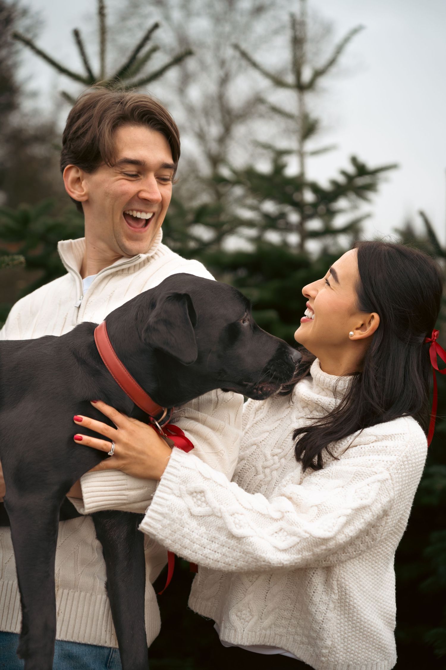 A couple in white sweaters share a joyful moment with their black dog outdoors among evergreen trees.