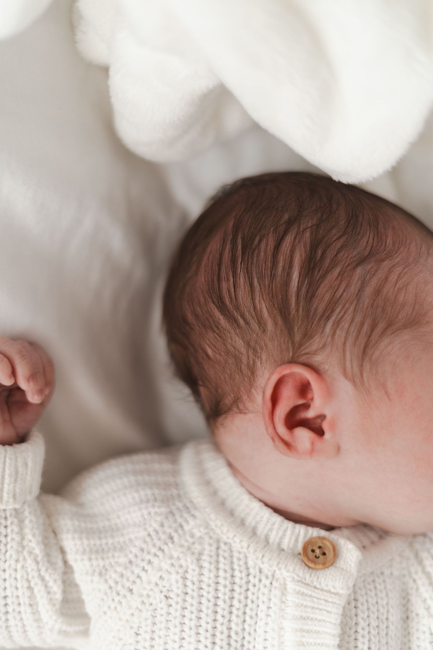 A sleeping newborn wearing a white knit cardigan rests peacefully against soft white bedding.
