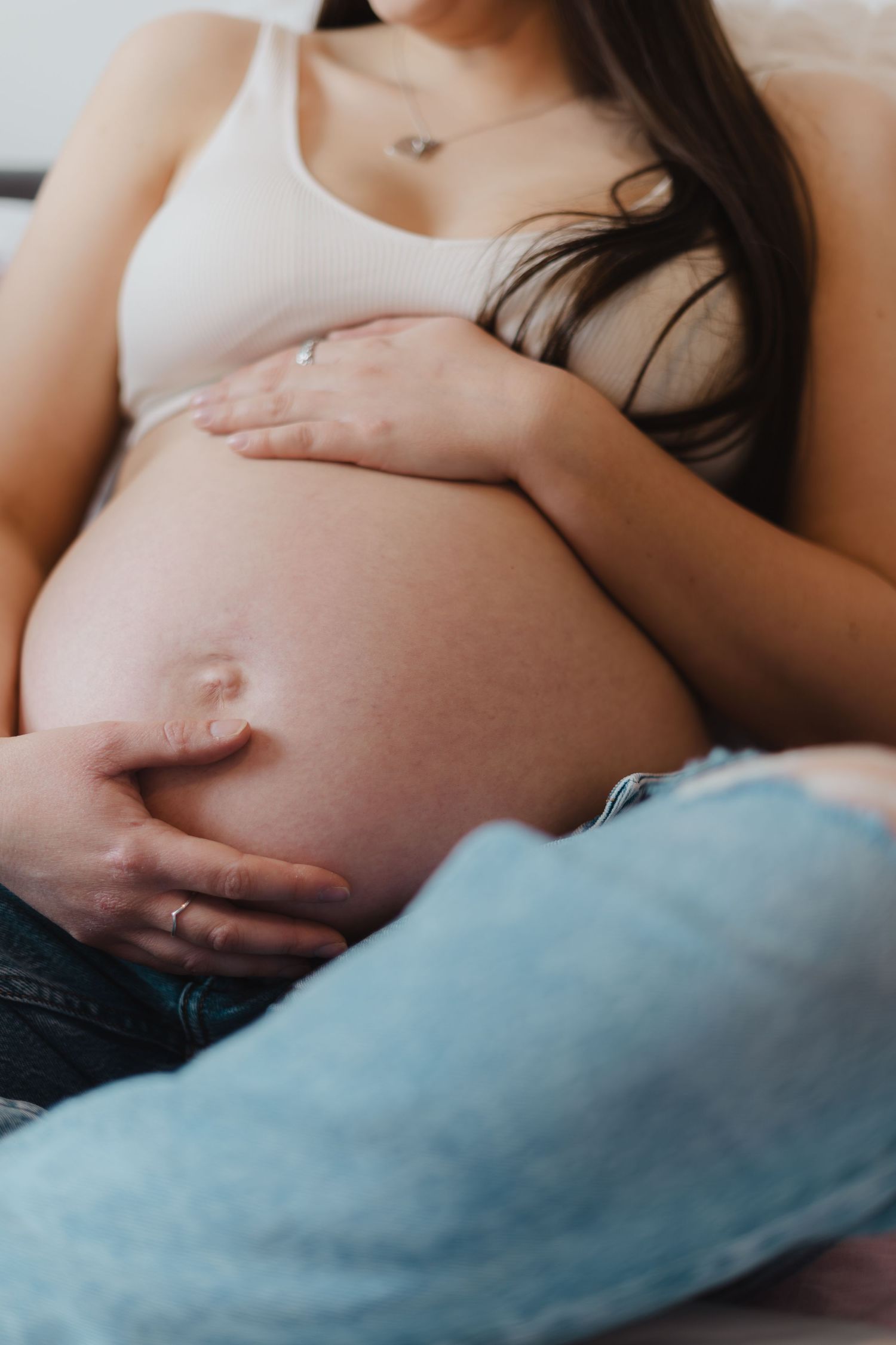 A pregnant woman in a white tank top gently cradles her bare belly while resting on blue bedding.