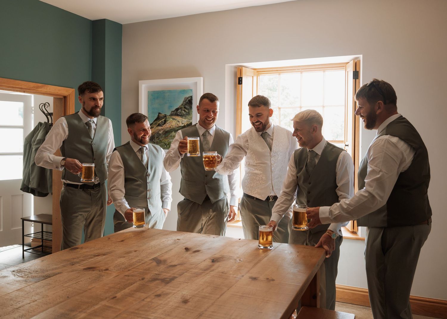 Groomsmen in gray vests toasting with beer mugs around a wooden table before a wedding ceremony.