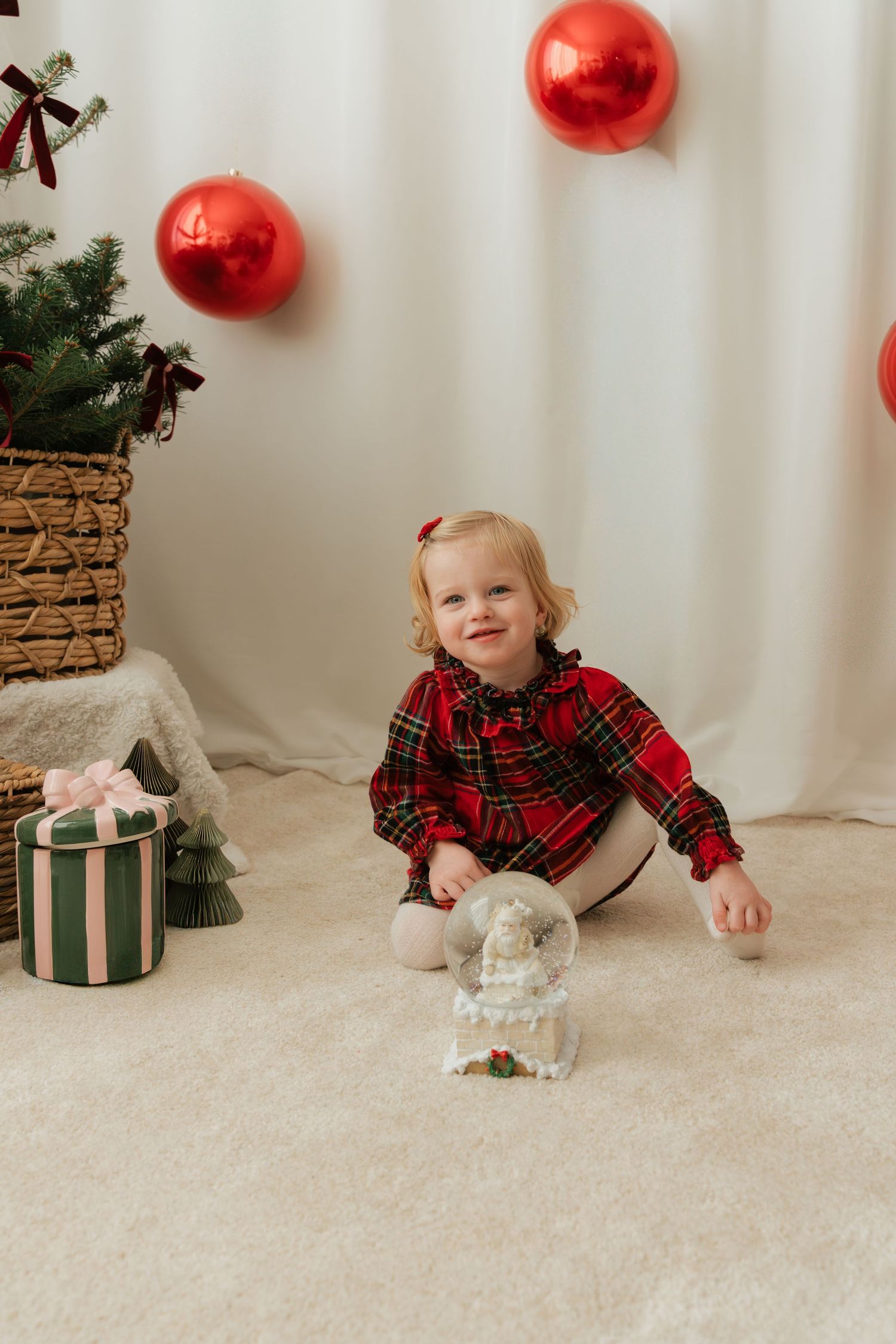 Toddler girl in red plaid dress sits on cream carpet near Christmas tree, gifts, and snow globe with red balloons.