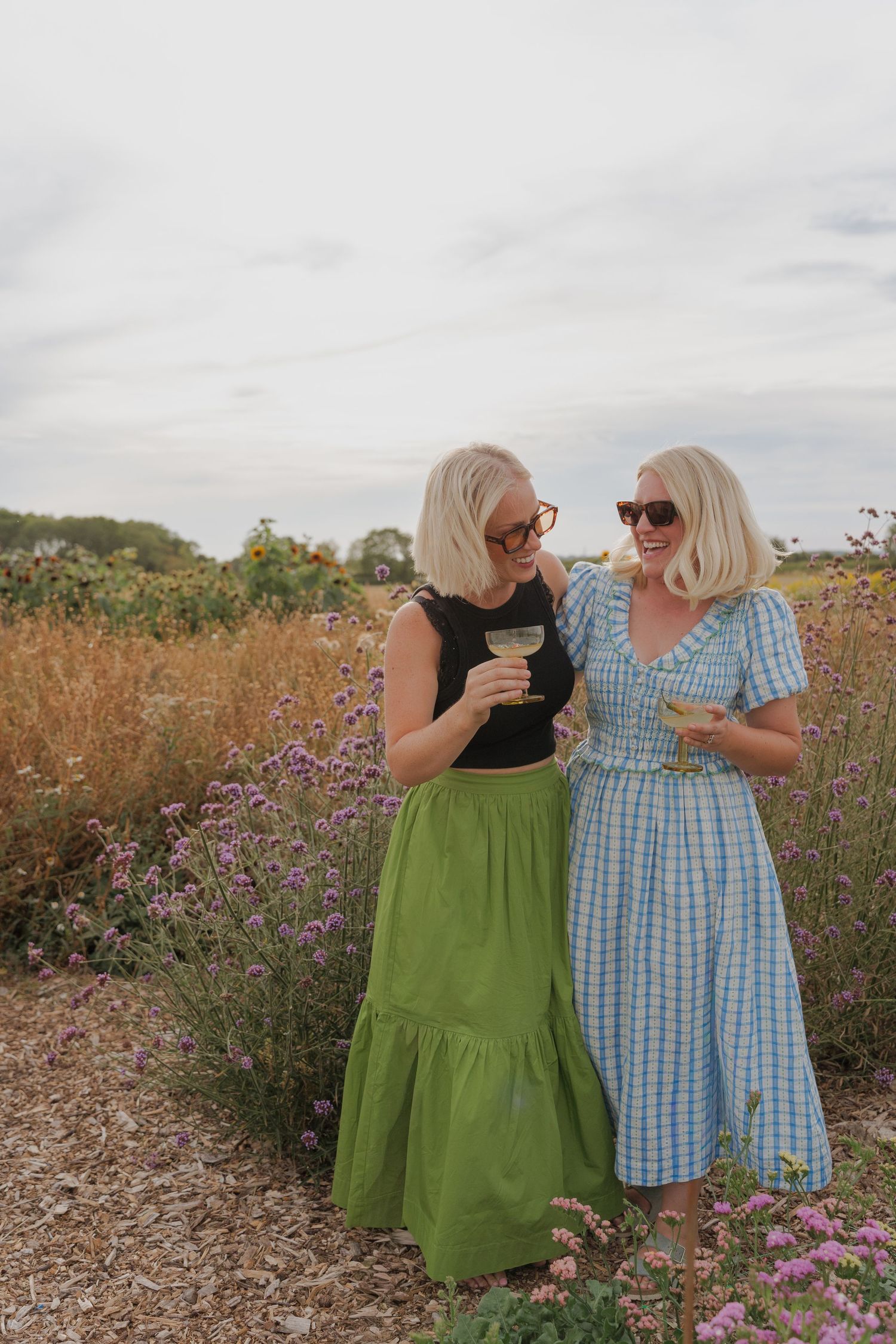 Two blonde women in sundresses holding cocktails laugh together in a wildflower field on a cloudy day.