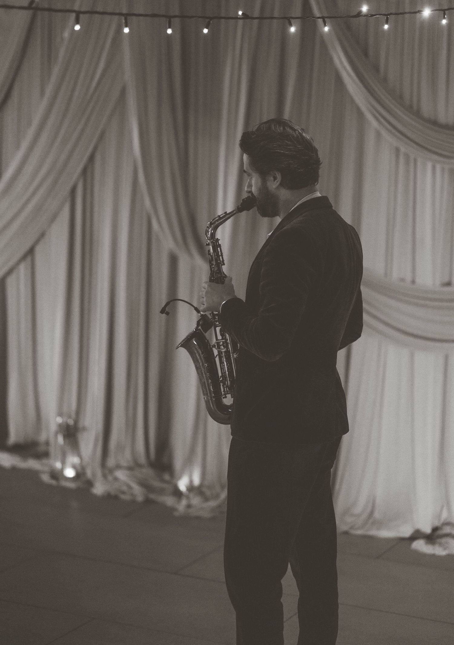 A man in a suit plays saxophone in front of elegant white drapes with string lights in a black and white photo.