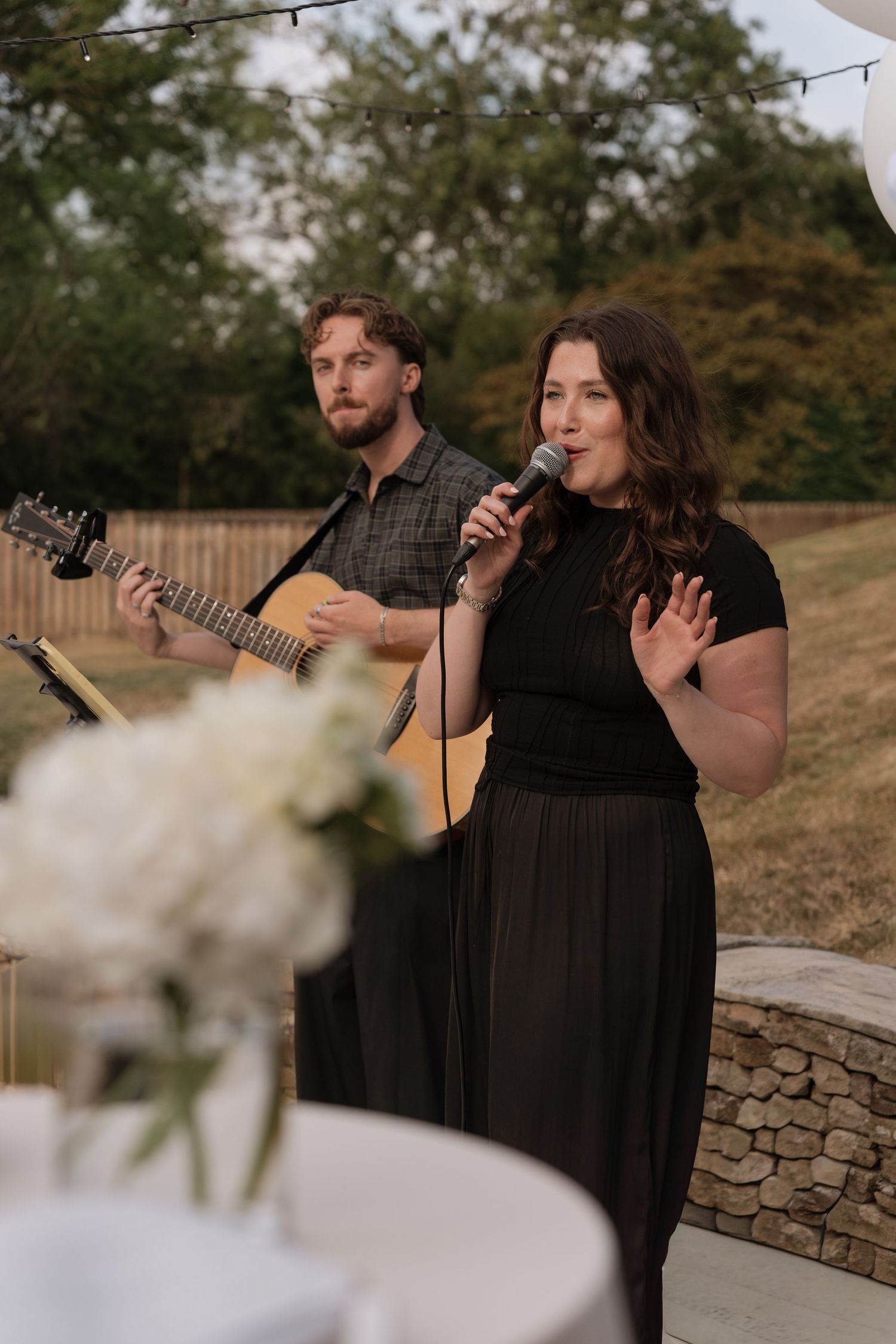 A woman sings into a microphone while a man plays acoustic guitar at an outdoor wedding reception.
