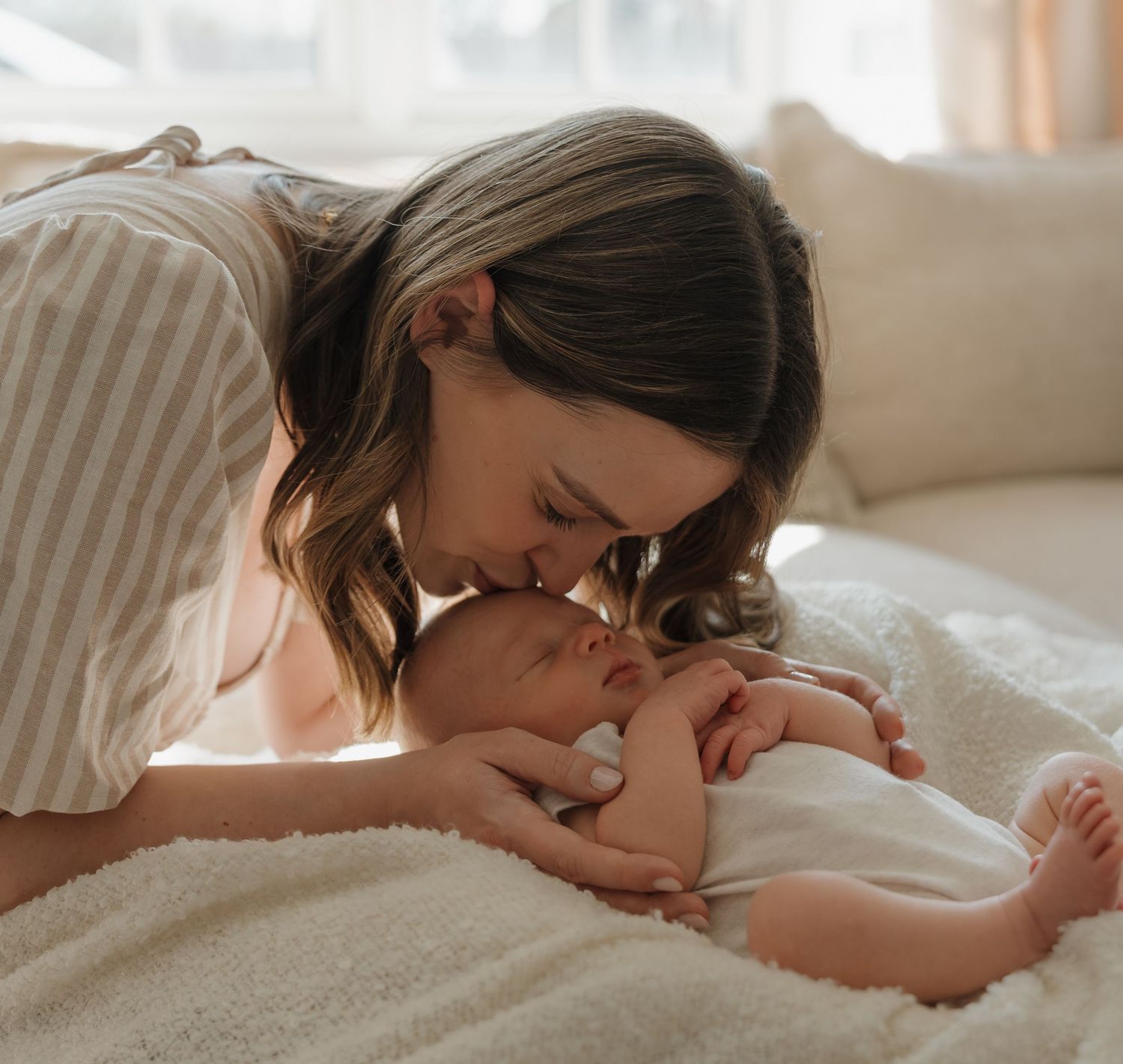 A tender mother gently kisses her sleeping newborn baby on a soft white blanket in a warmly lit room.