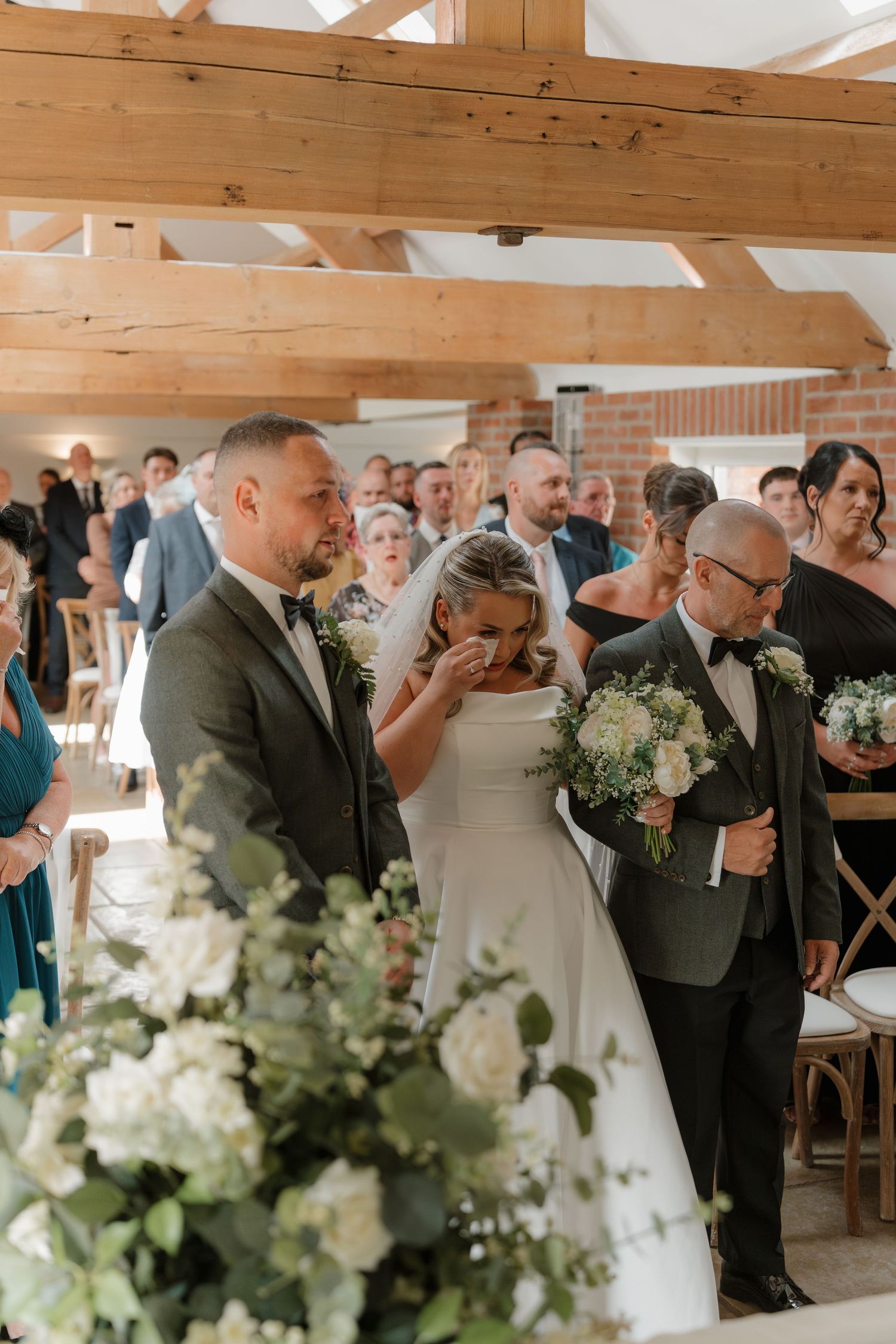 Emotional bride walks down the aisle with her father in a rustic barn wedding venue, guests watching in background.