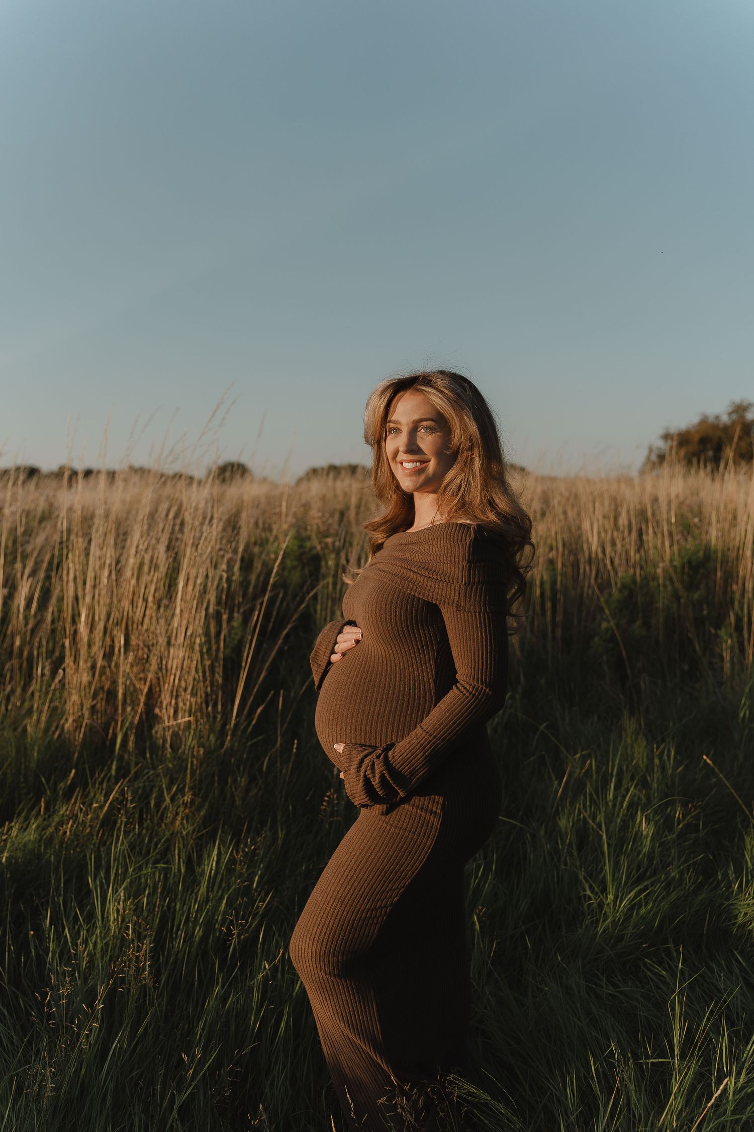 A maternity photo series in a golden field shows a person in a brown dress posing against tall grass at sunset.