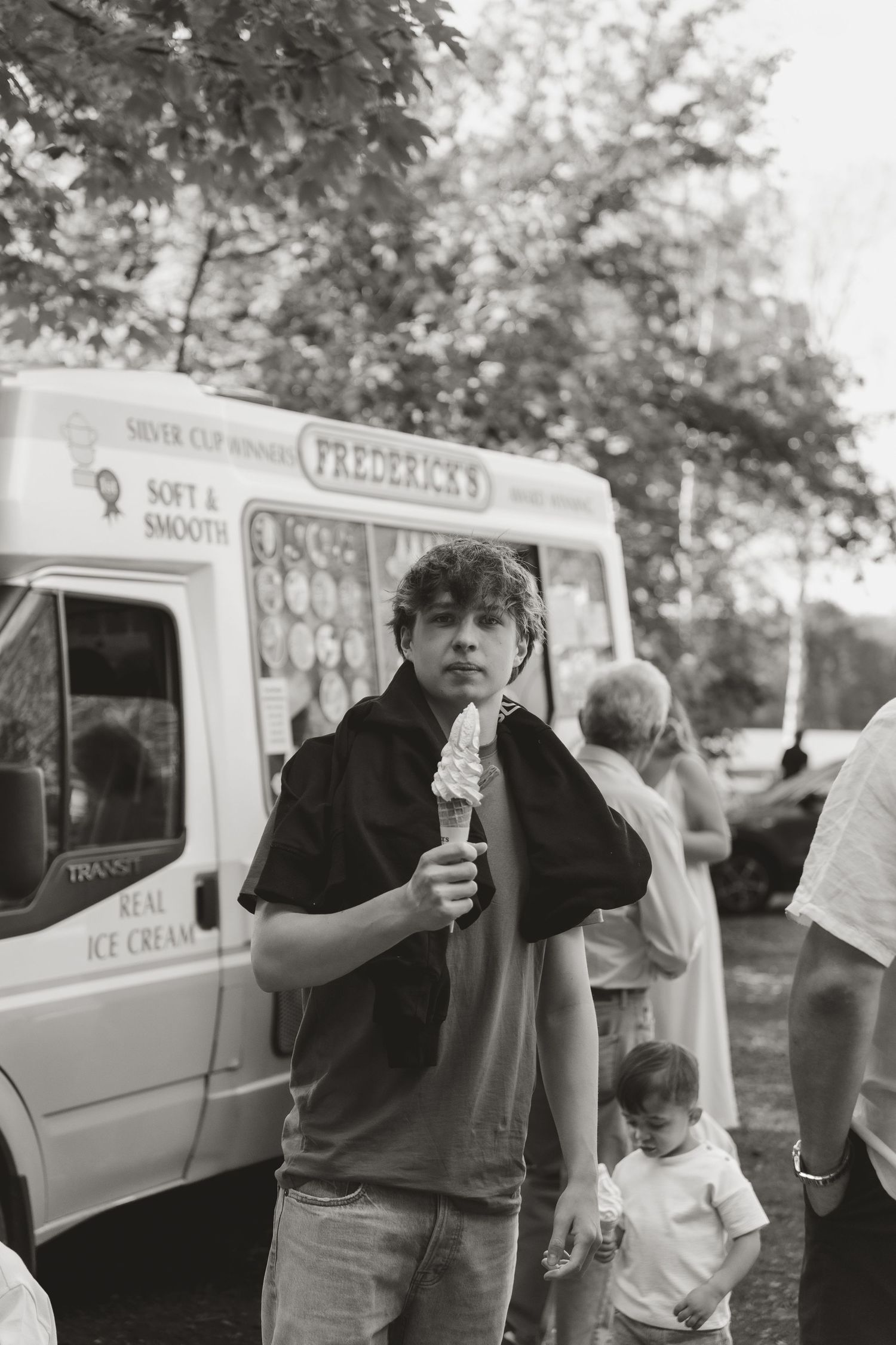Young man eating ice cream cone in black and white photo near Frederick's ice cream van outdoors.