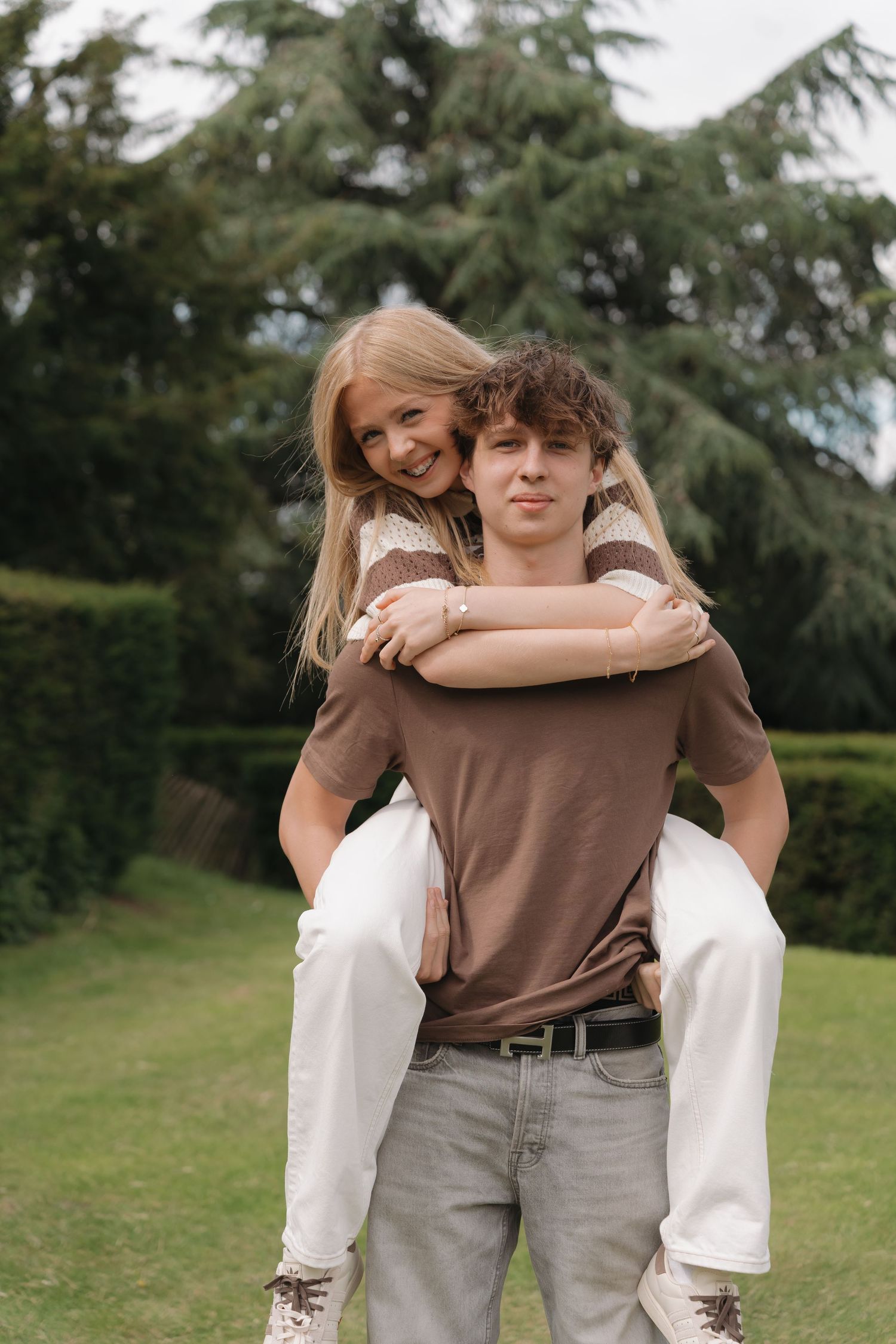 Young couple in a park, man giving woman a piggyback ride, both smiling, surrounded by green trees.