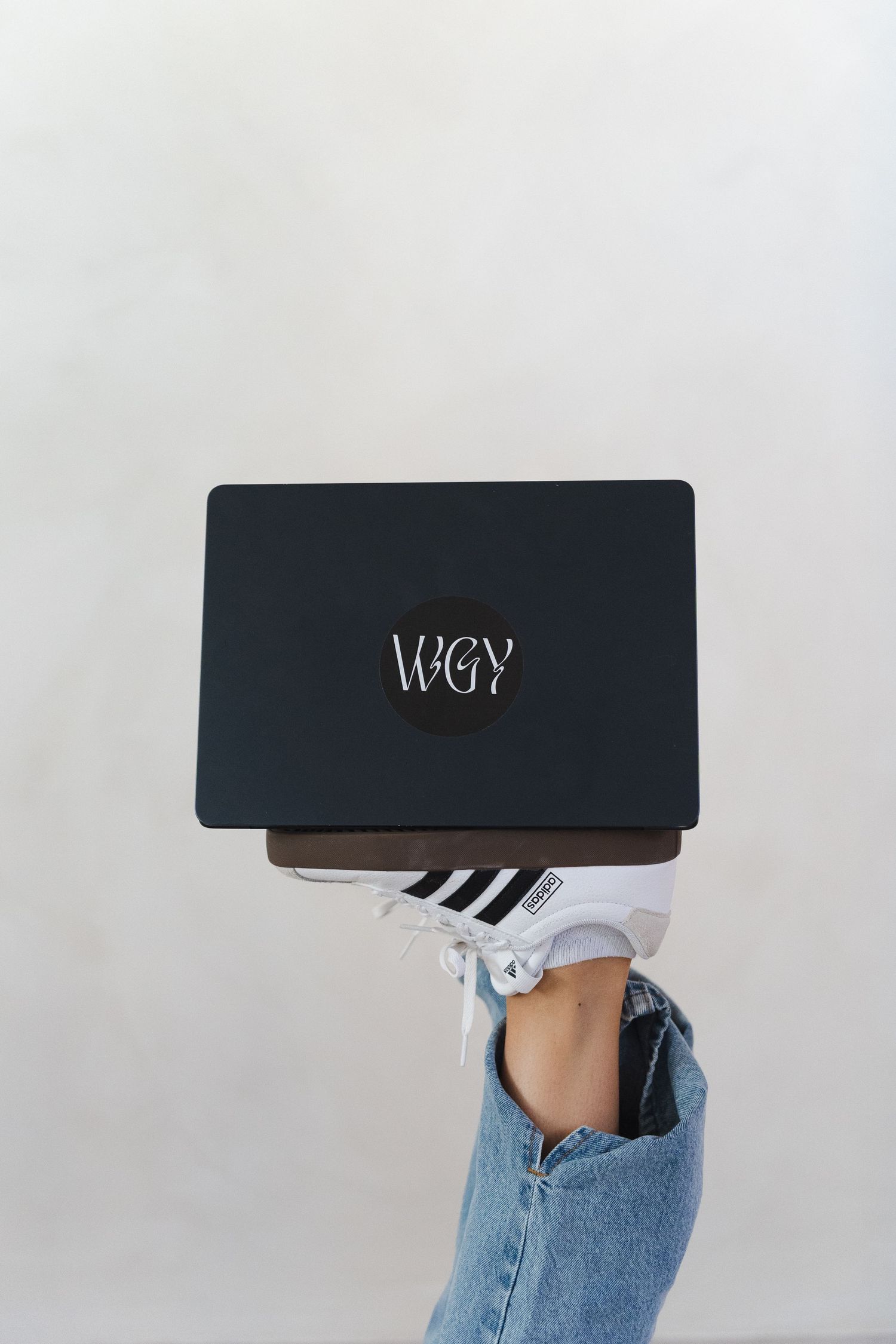 A black laptop with VEGY branding balanced on someone's knee wearing blue jeans and white sneakers.