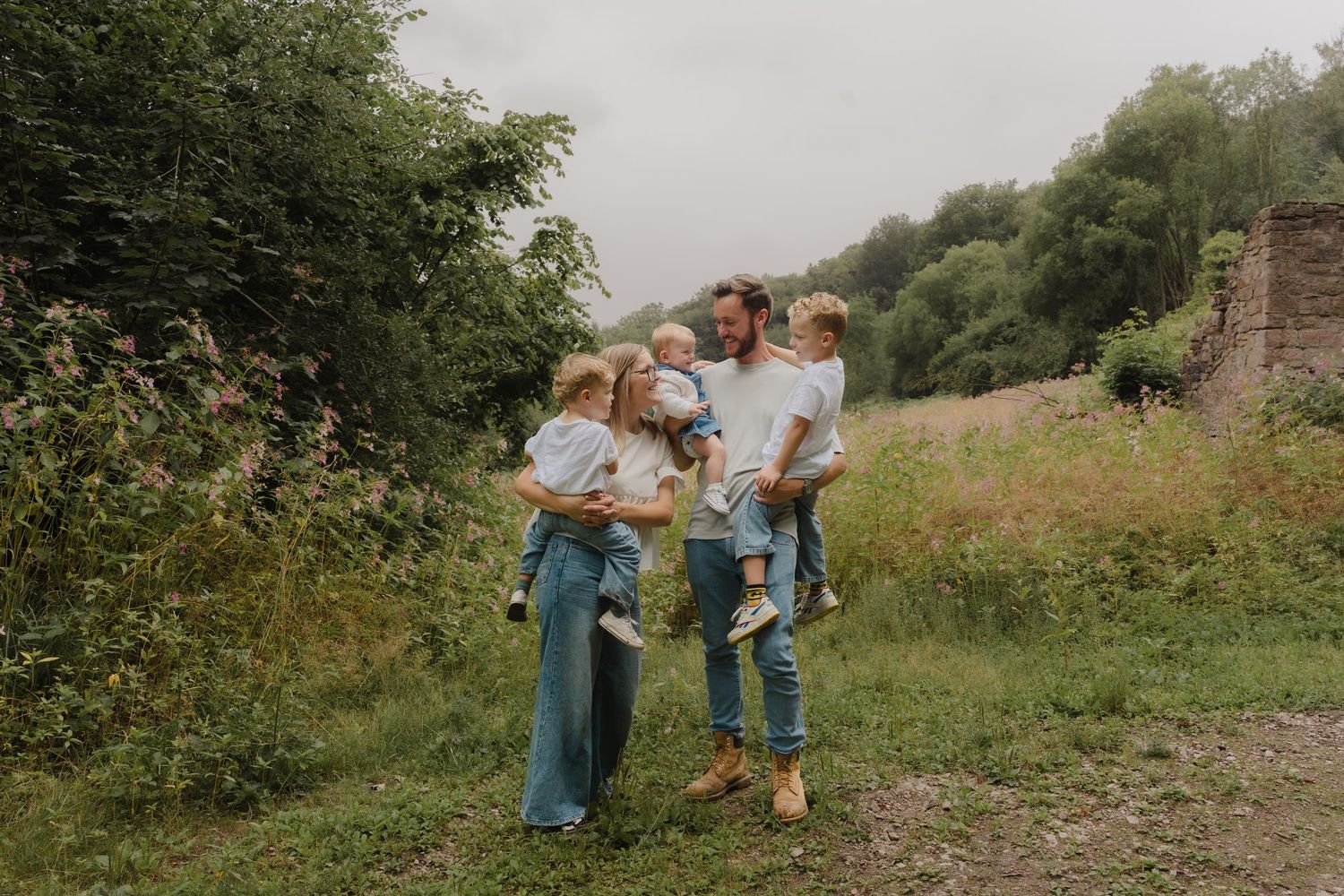 A couple in rustic outdoor attire embraces while standing in a lush green field on a cloudy day.