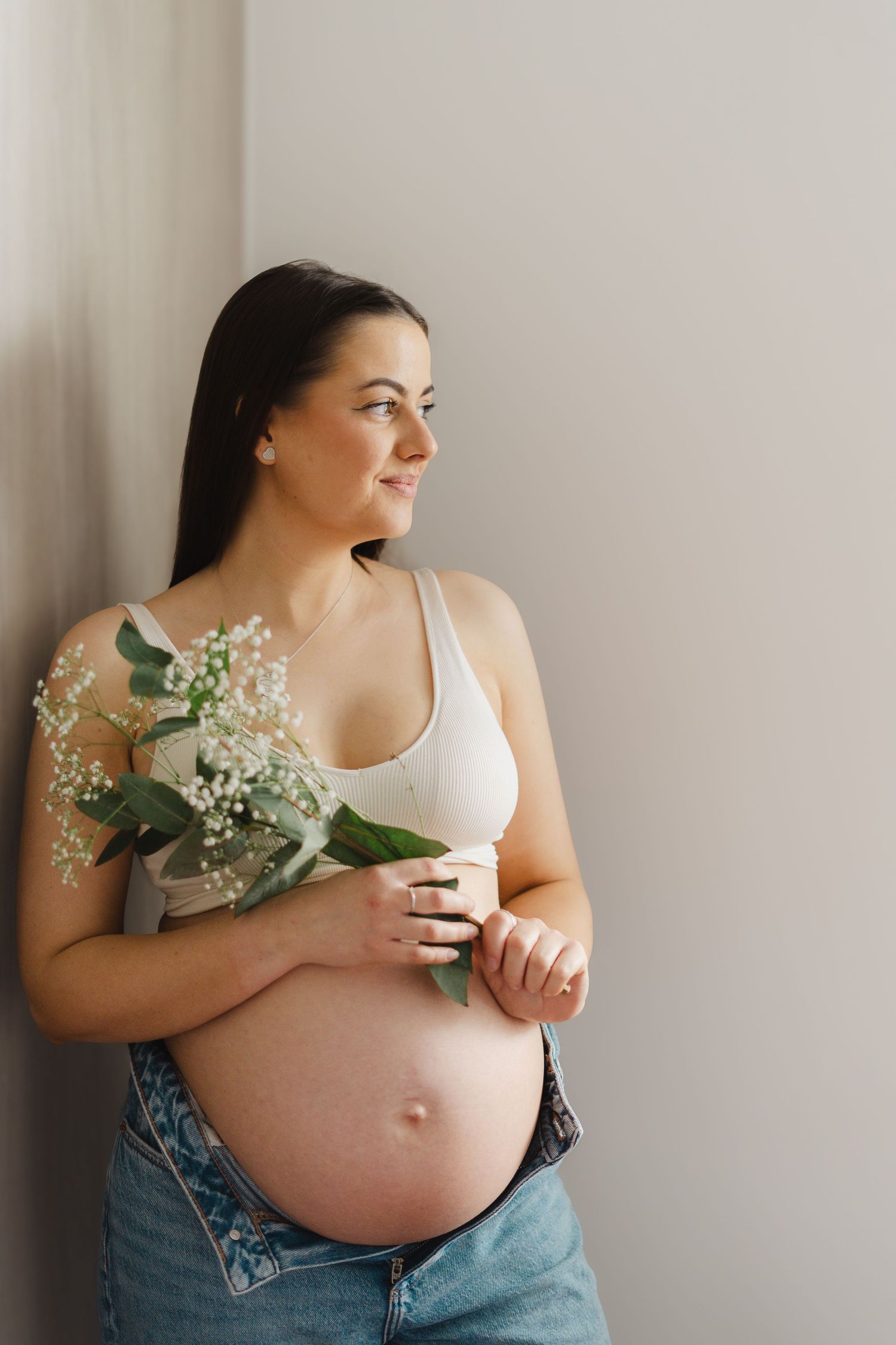 A pregnant person in a white top and jeans holds delicate white flowers while posing against a light wall in a photo series.