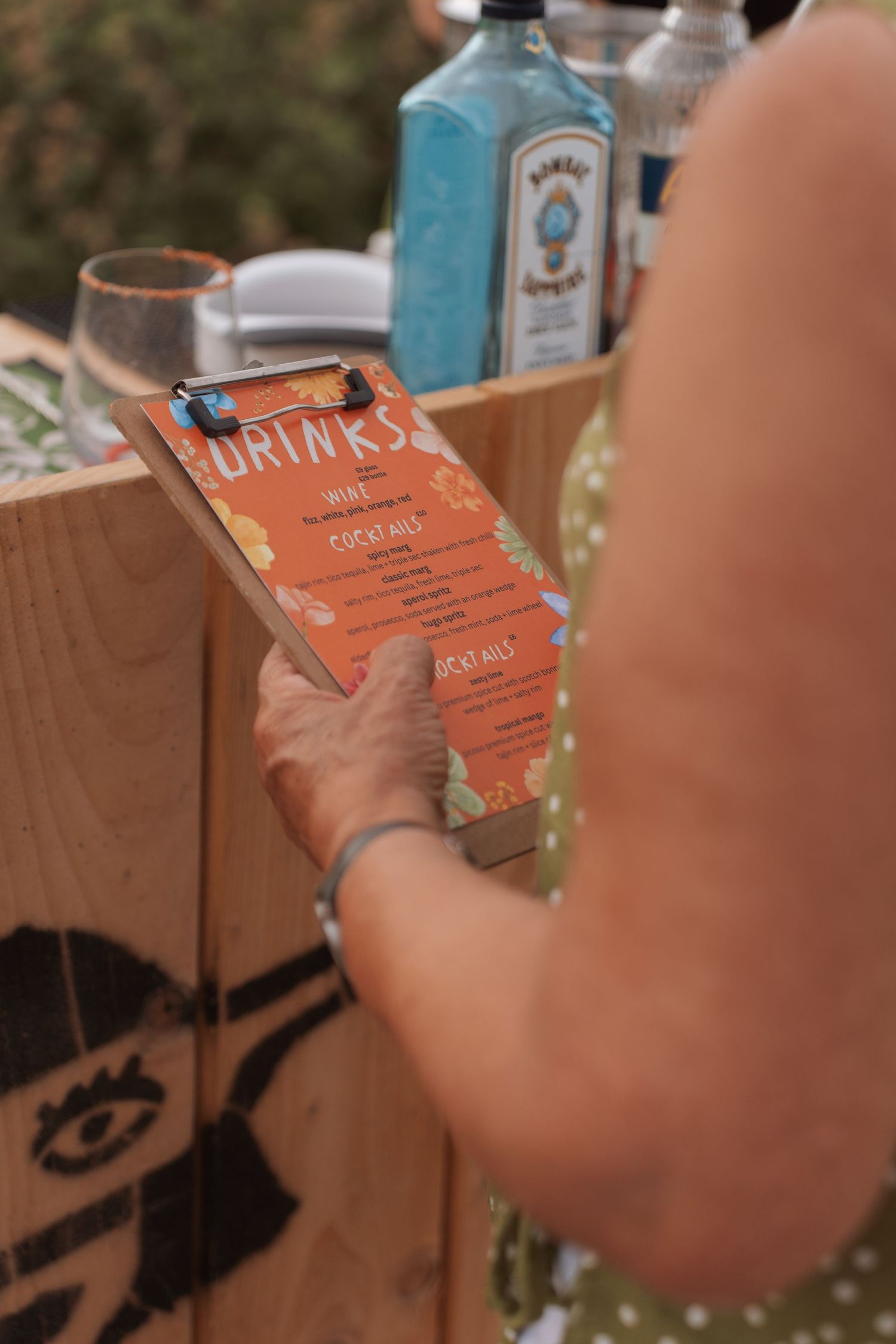 Person holding a clipboard with an orange drinks menu at an outdoor bar with Bombay Sapphire gin bottles.