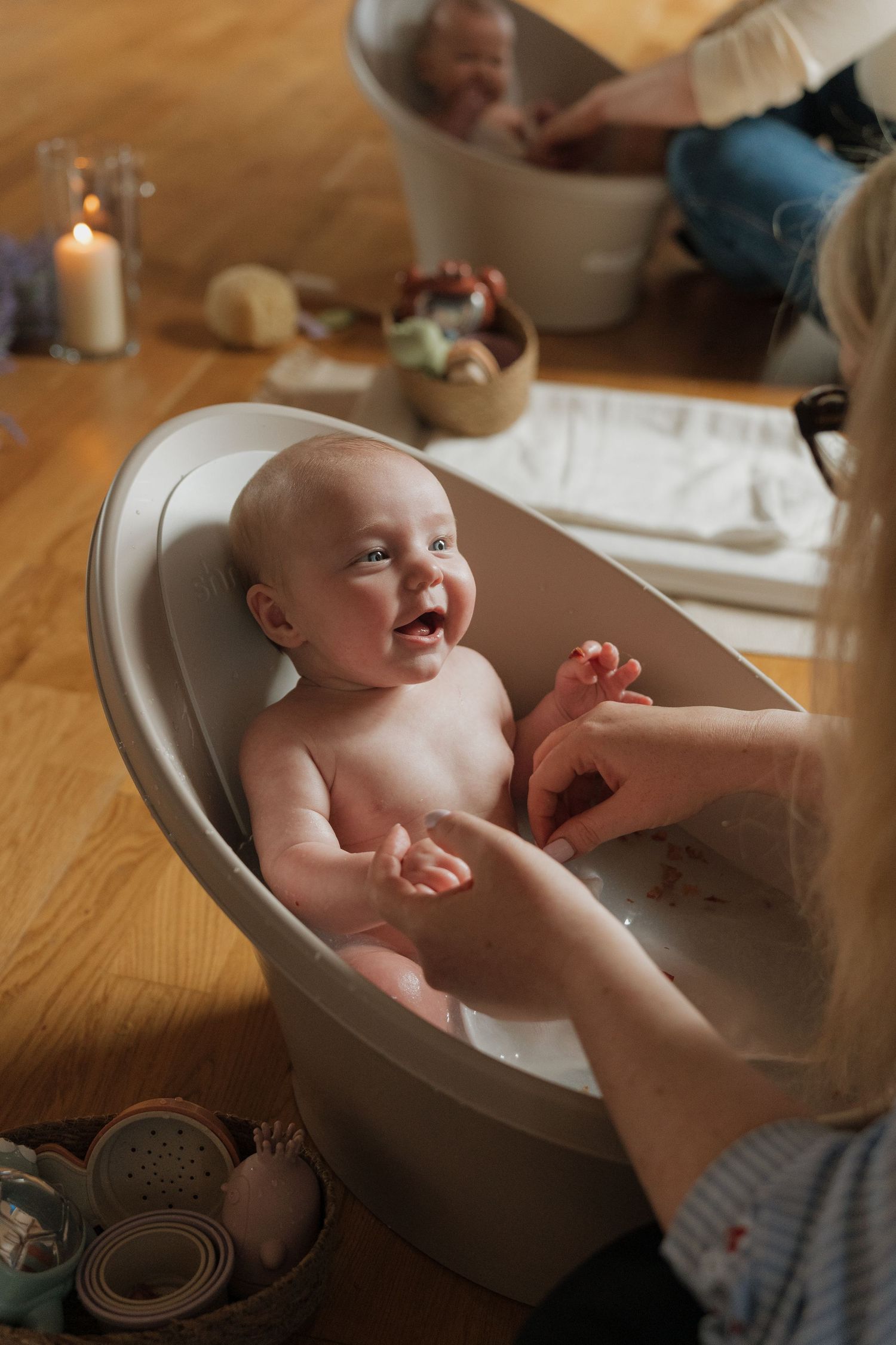 Happy baby smiling in a small white infant bathtub on a wooden floor while a caregiver gently holds their feet.