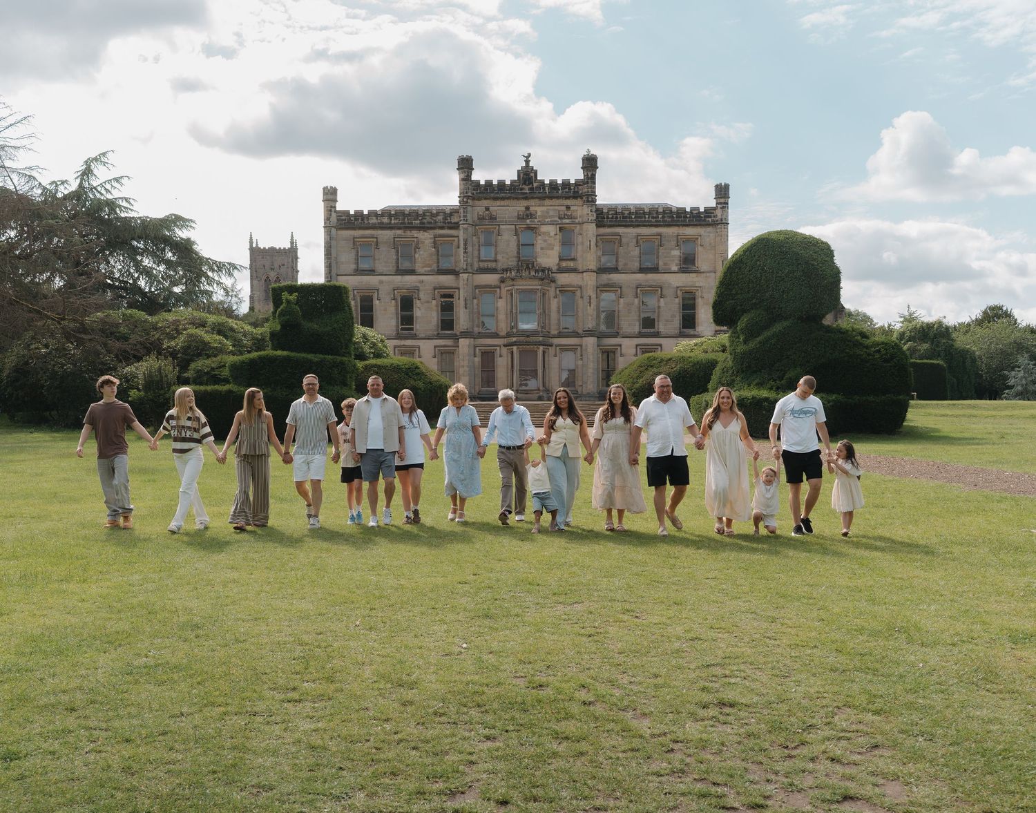 Large family group holding hands on lawn before grand historic English manor house on cloudy day.