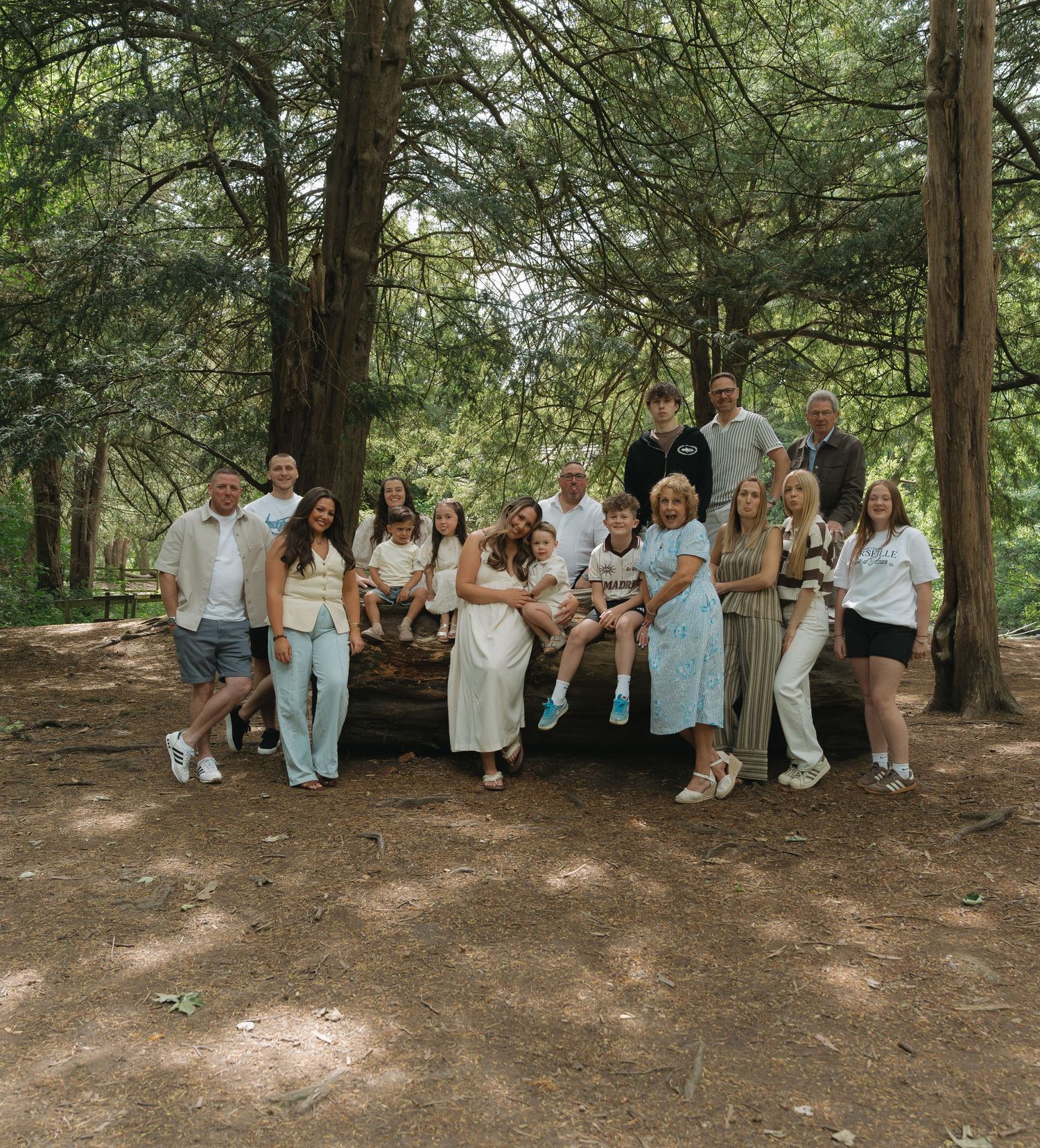Large family group photo taken outdoors in a pine forest, posing together near a fallen log in natural light.