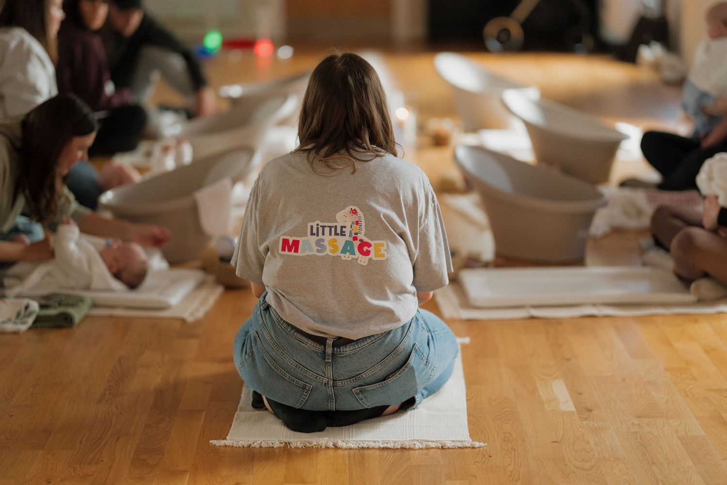 Parents and babies attend a Little Massage class, sitting on mats in a circle on a wooden floor.