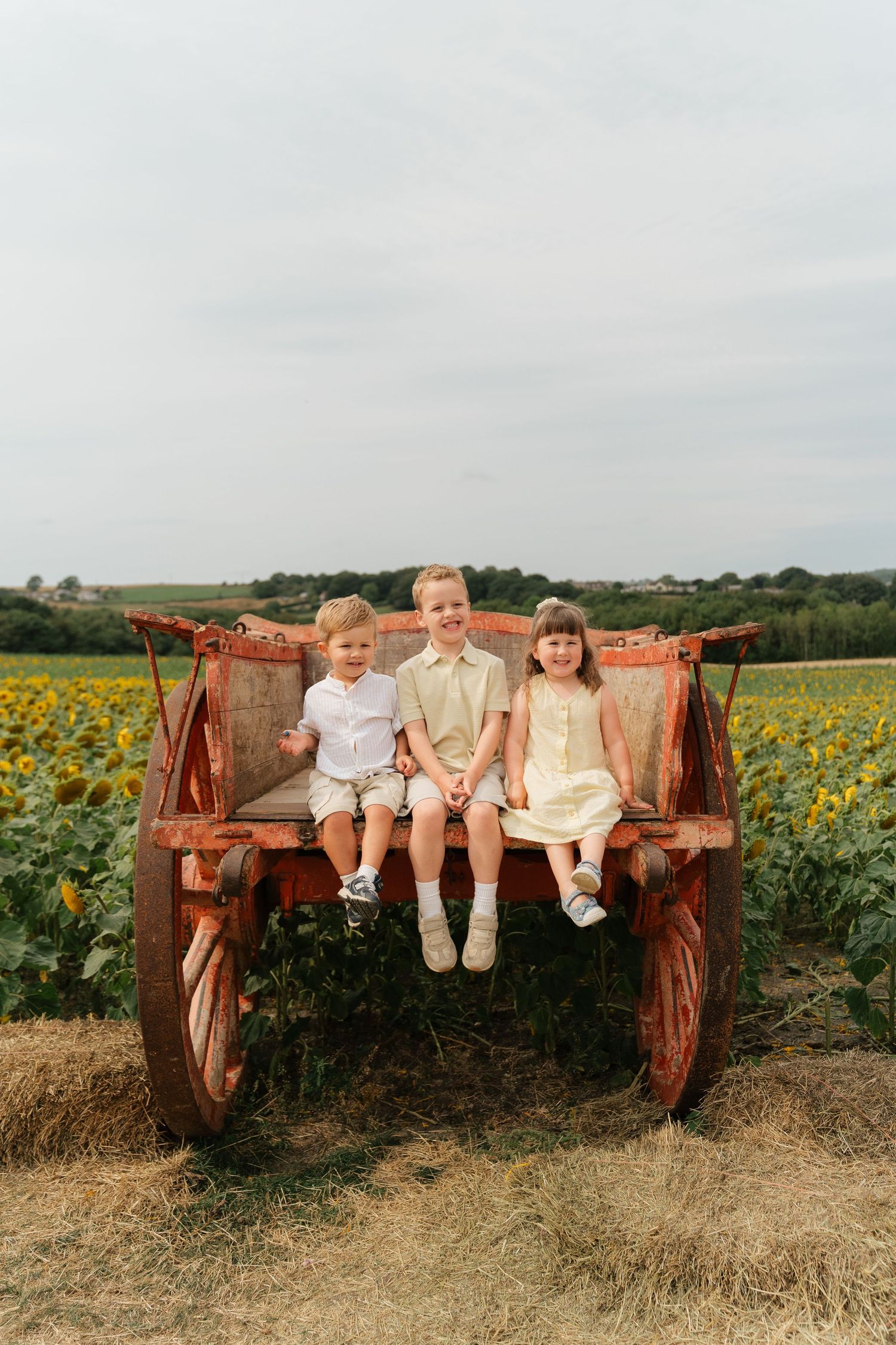 Three children sit smiling on a rustic wooden wagon in front of a sunflower field on a cloudy day.