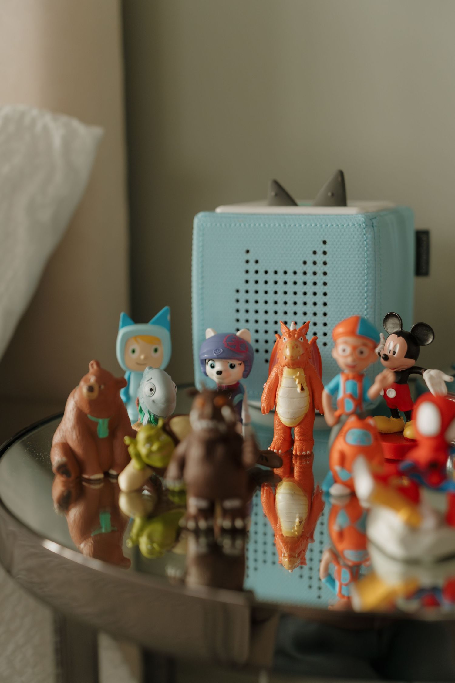Colorful toy figures including Mickey Mouse arranged on a glass table beside a teal Toniebox children's music player.