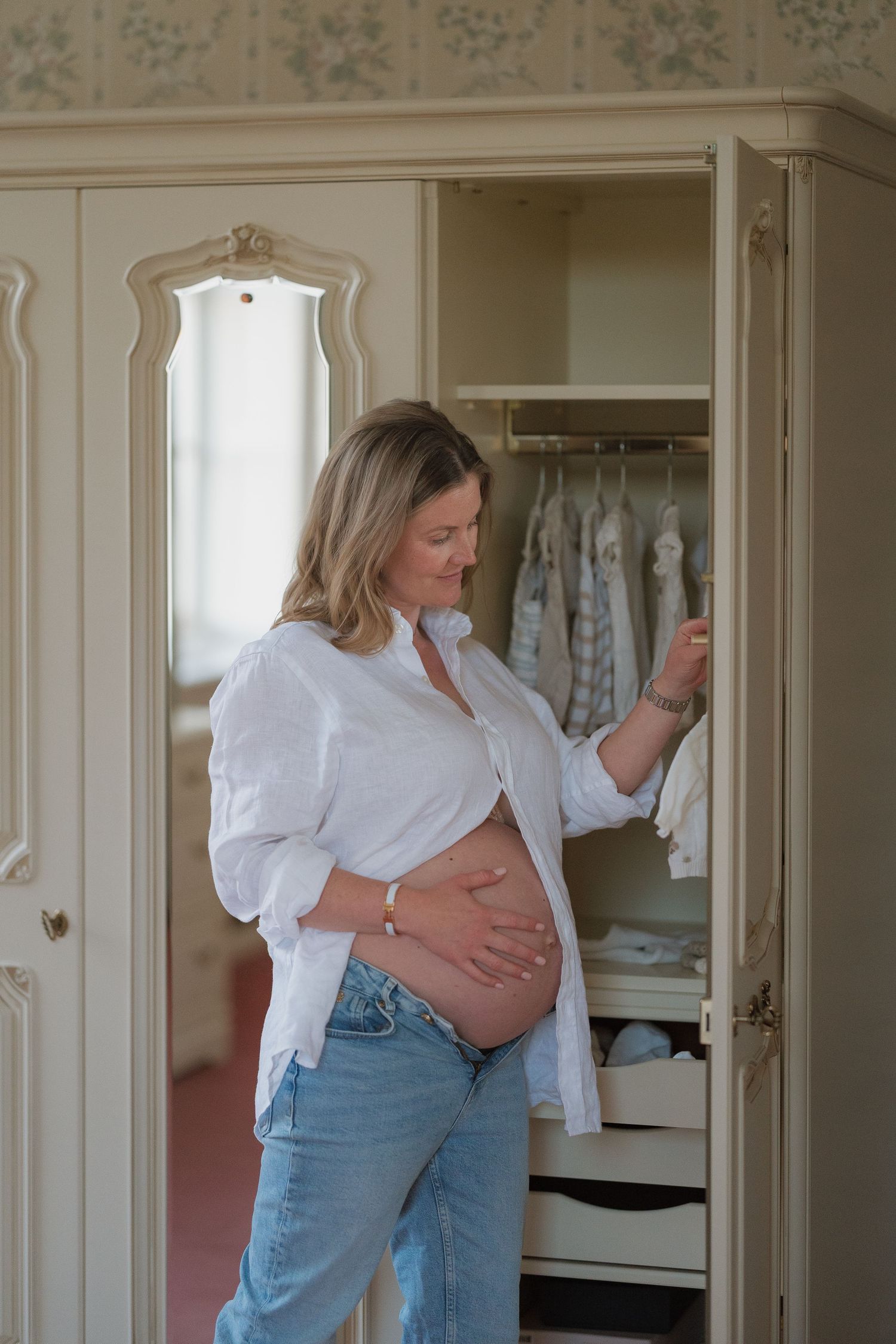 Pregnant woman in white shirt and jeans holding her baby bump while browsing a wardrobe of newborn clothes.