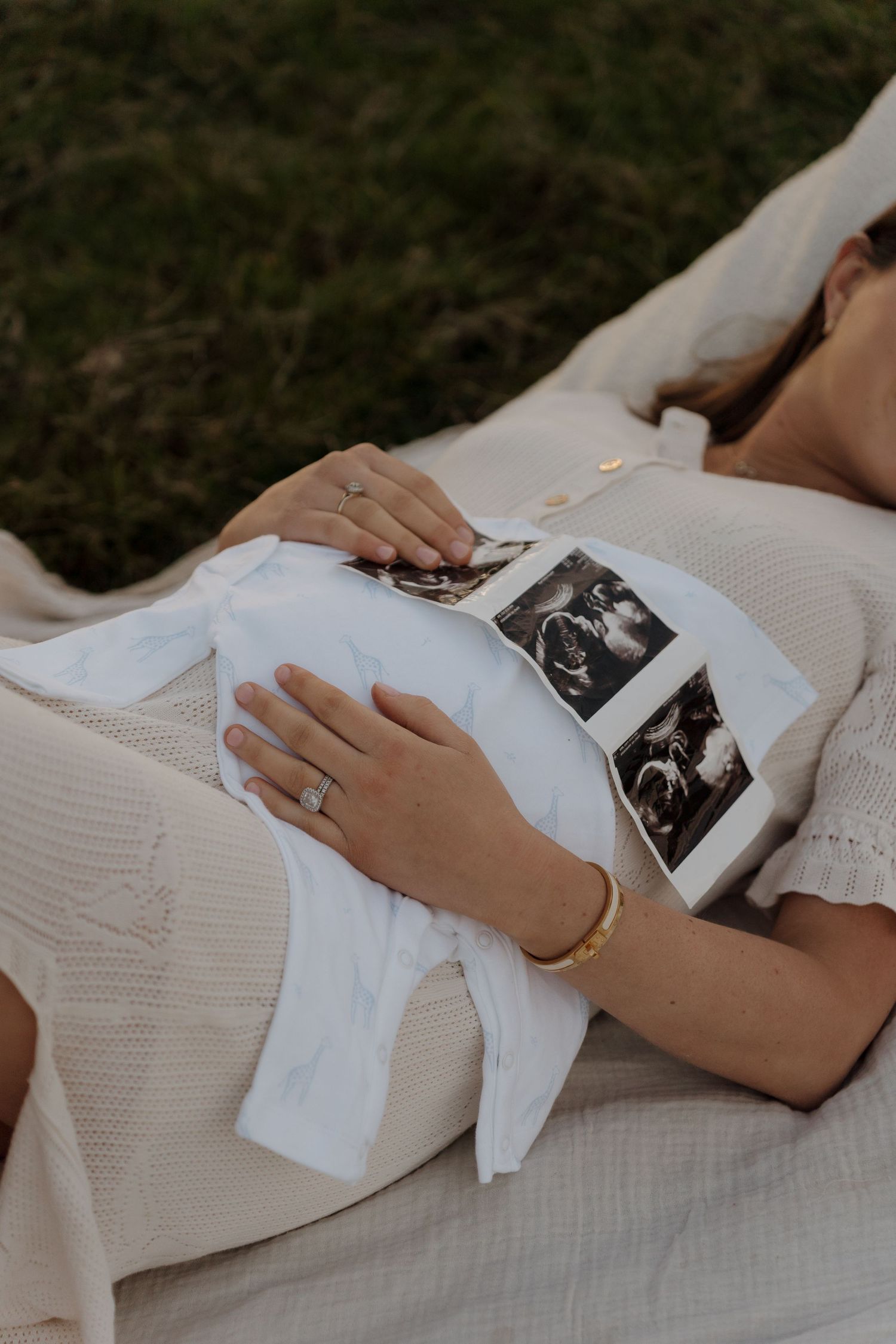 A person in a white dress holds ultrasound photos while resting on a blanket outdoors.