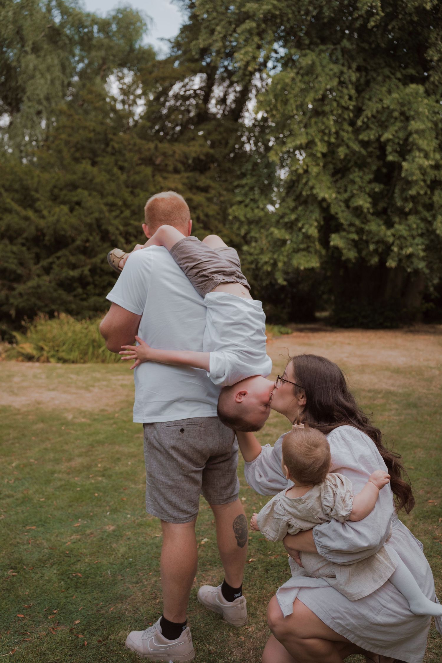 A family of four shares a loving moment outdoors, with dad lifting a child as mom kisses them while holding a baby.