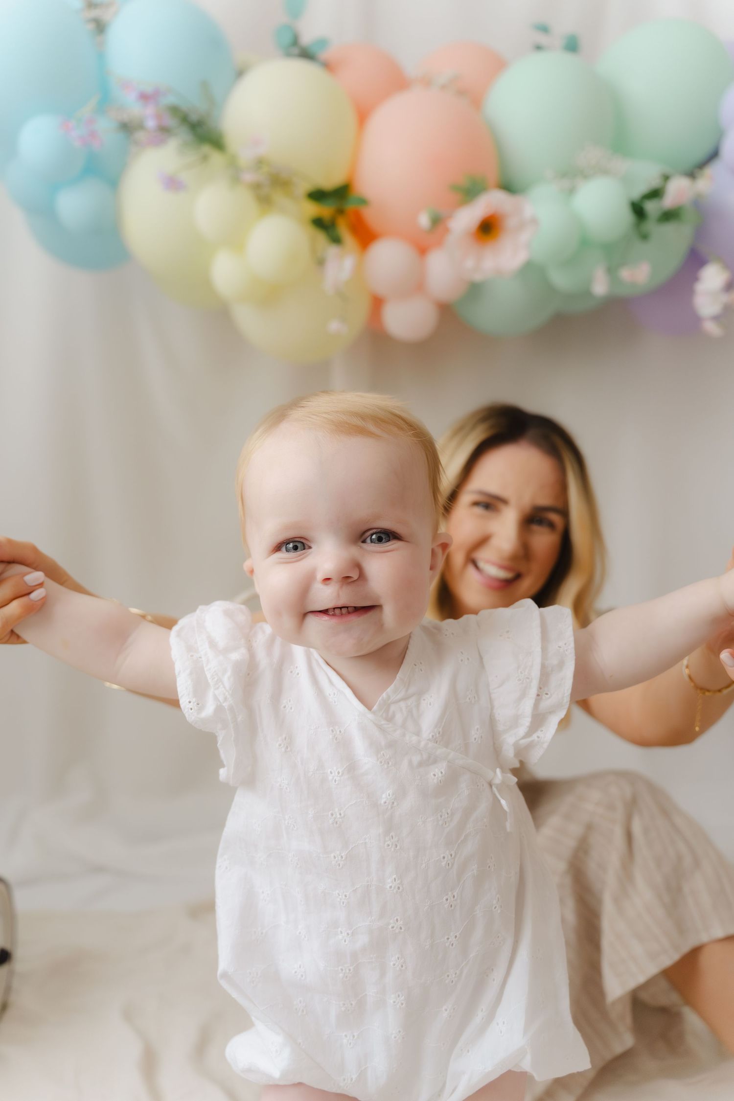 Pastel balloon garland decorates a photo session featuring a baby in white dress against light backdrop.